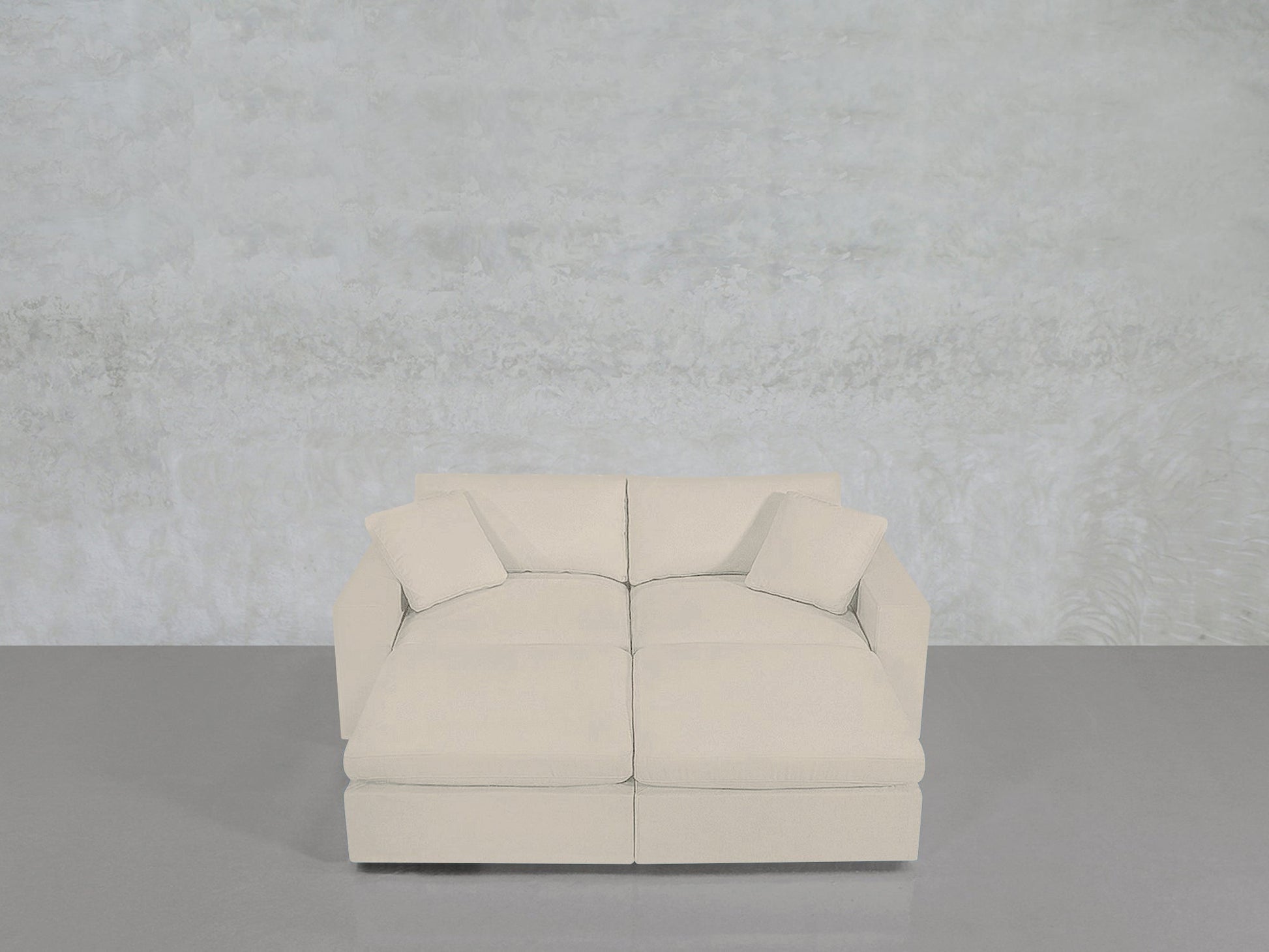 A light beige, modern 4-Seat Modular Daybed with two matching throw pillows sits on a smooth gray floor against a textured pale gray wall.