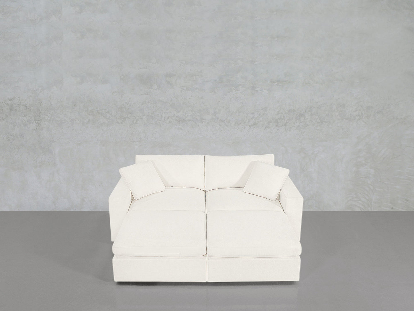 A 4-Seat Modular Daybed with a modern white design and two matching pillows sits on a smooth gray floor against a light gray textured wall.