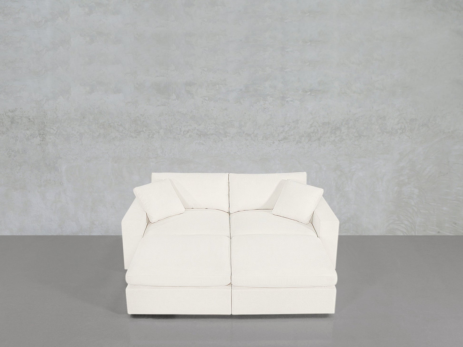 A 4-Seat Modular Daybed with a modern white design and two matching pillows sits on a smooth gray floor against a light gray textured wall.