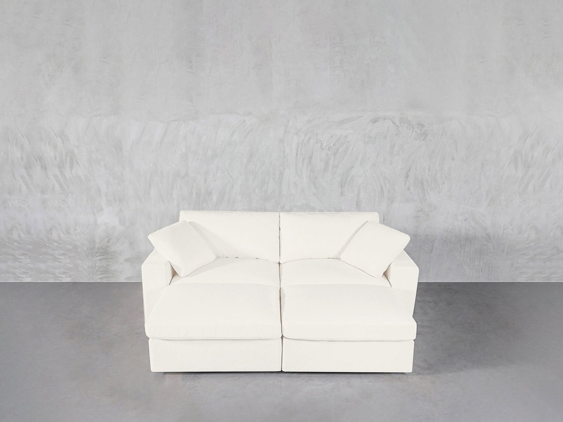 A modern white 4-Seat Modular Daybed with two seat cushions and two matching pillows sits against a textured light gray wall on a smooth gray floor.