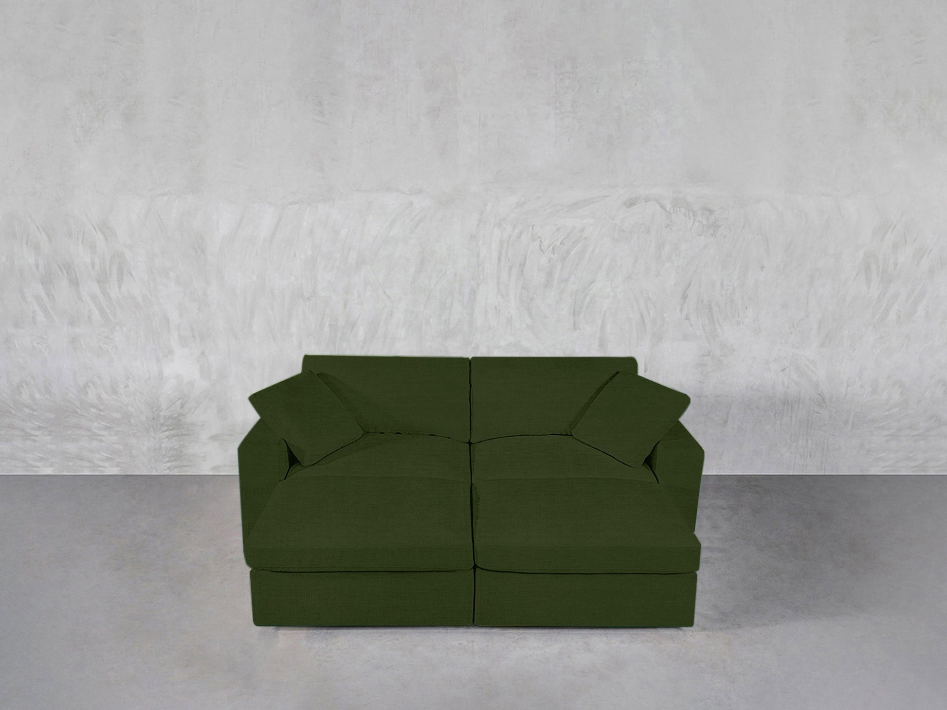 A 4-Seat Modular Daybed in dark green with two matching cushions rests on a smooth, light gray floor against a textured, light gray wall.