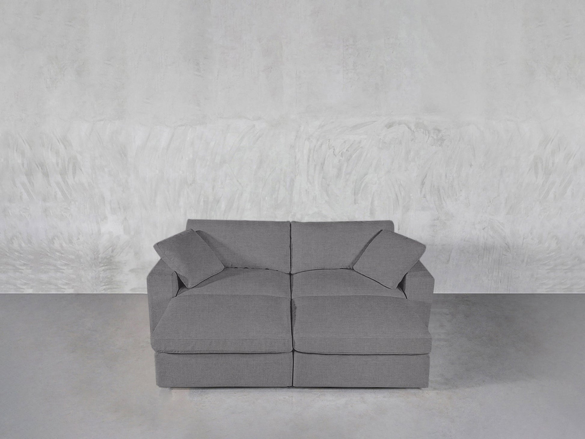 A 4-Seat Modular Daybed in modern gray with two seat cushions and two matching throw pillows is arranged against a textured light gray wall on a smooth gray floor.