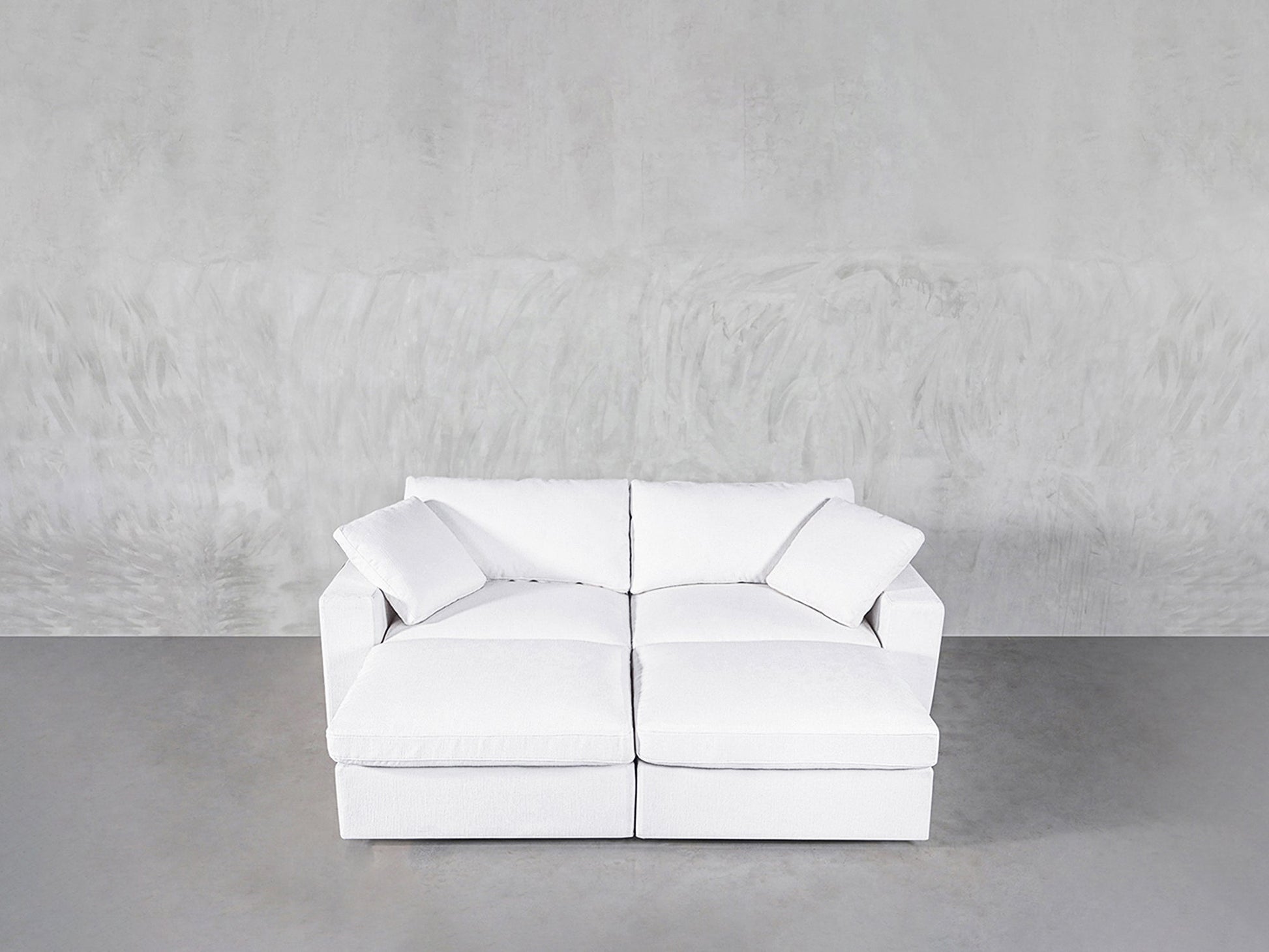 A modern white 4-Seat Modular Daybed with two seat and back cushions sits against a light gray textured wall on a smooth gray floor. Two matching white throw pillows are placed in each corner of the daybed.