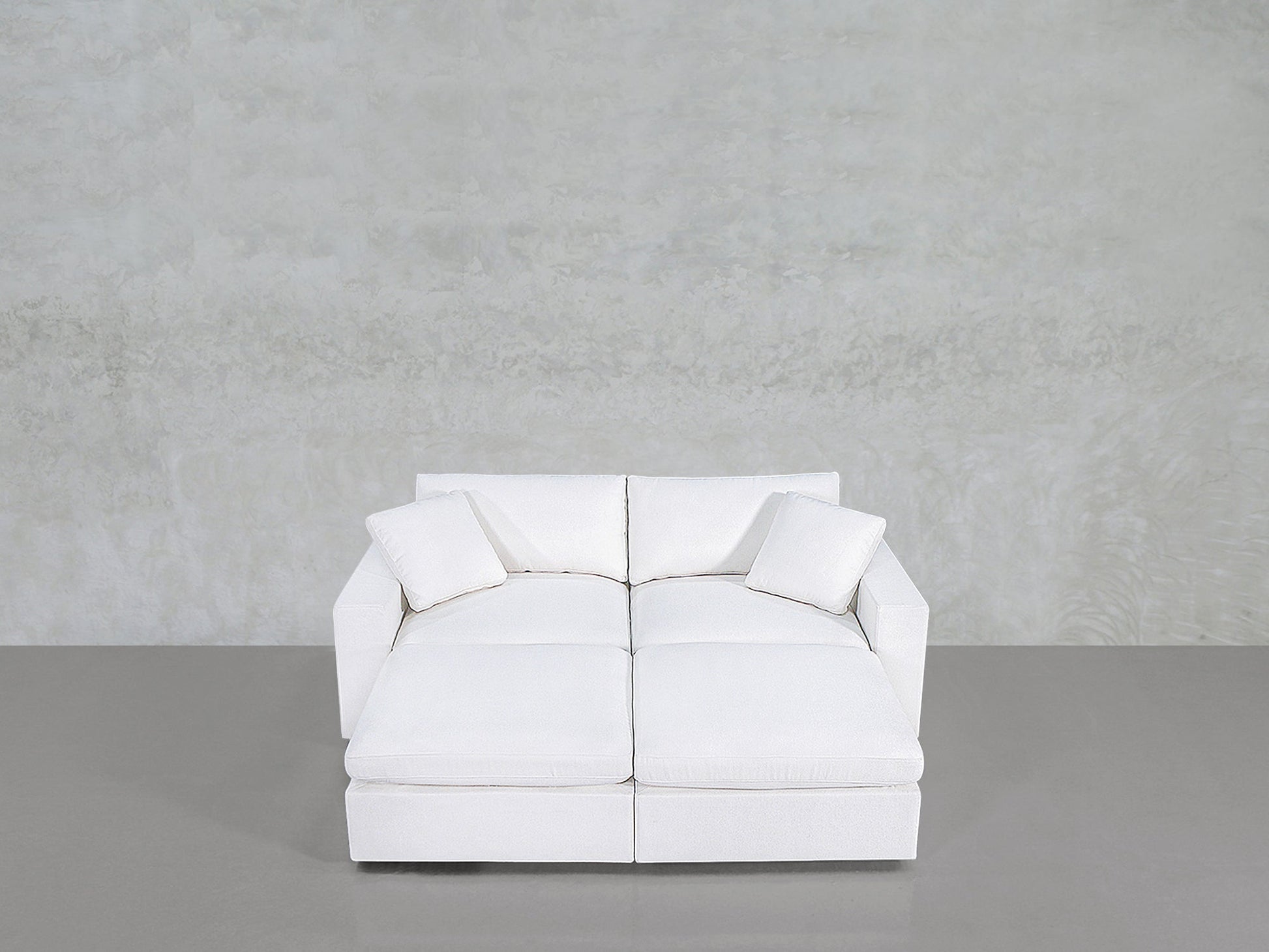 A modern, white 4-Seat Modular Daybed with two matching white cushions sits against a plain, light gray textured wall and a smooth gray floor.