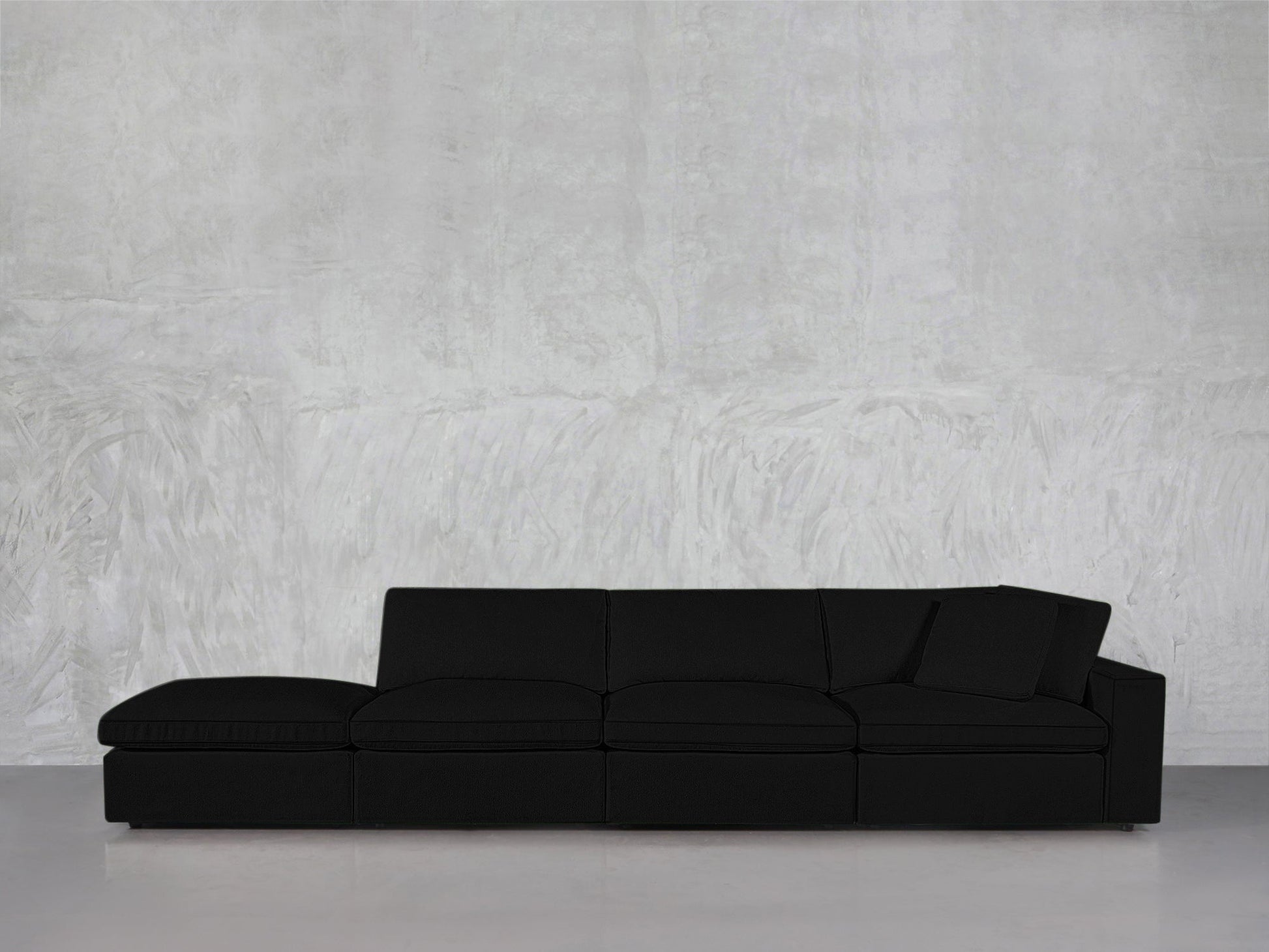 The 4-Seat Modular Lounger Sofa in black, featuring one armrest, sits against a light gray textured wall and rests on a smooth, light-colored floor.