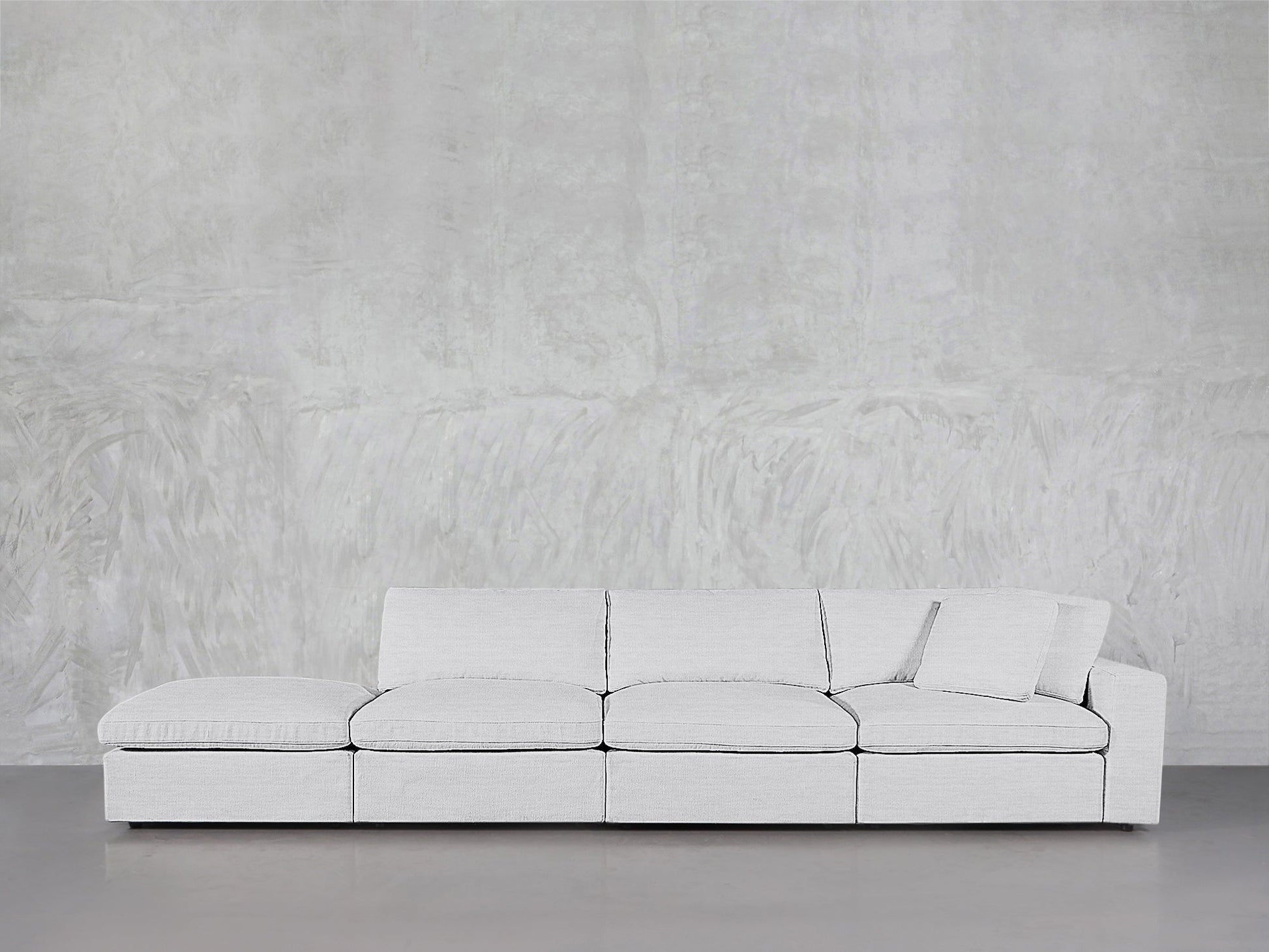 A modern 4-Seat Modular Lounger Sofa in light gray with matching pillow is set against a textured light gray wall on a polished concrete floor.