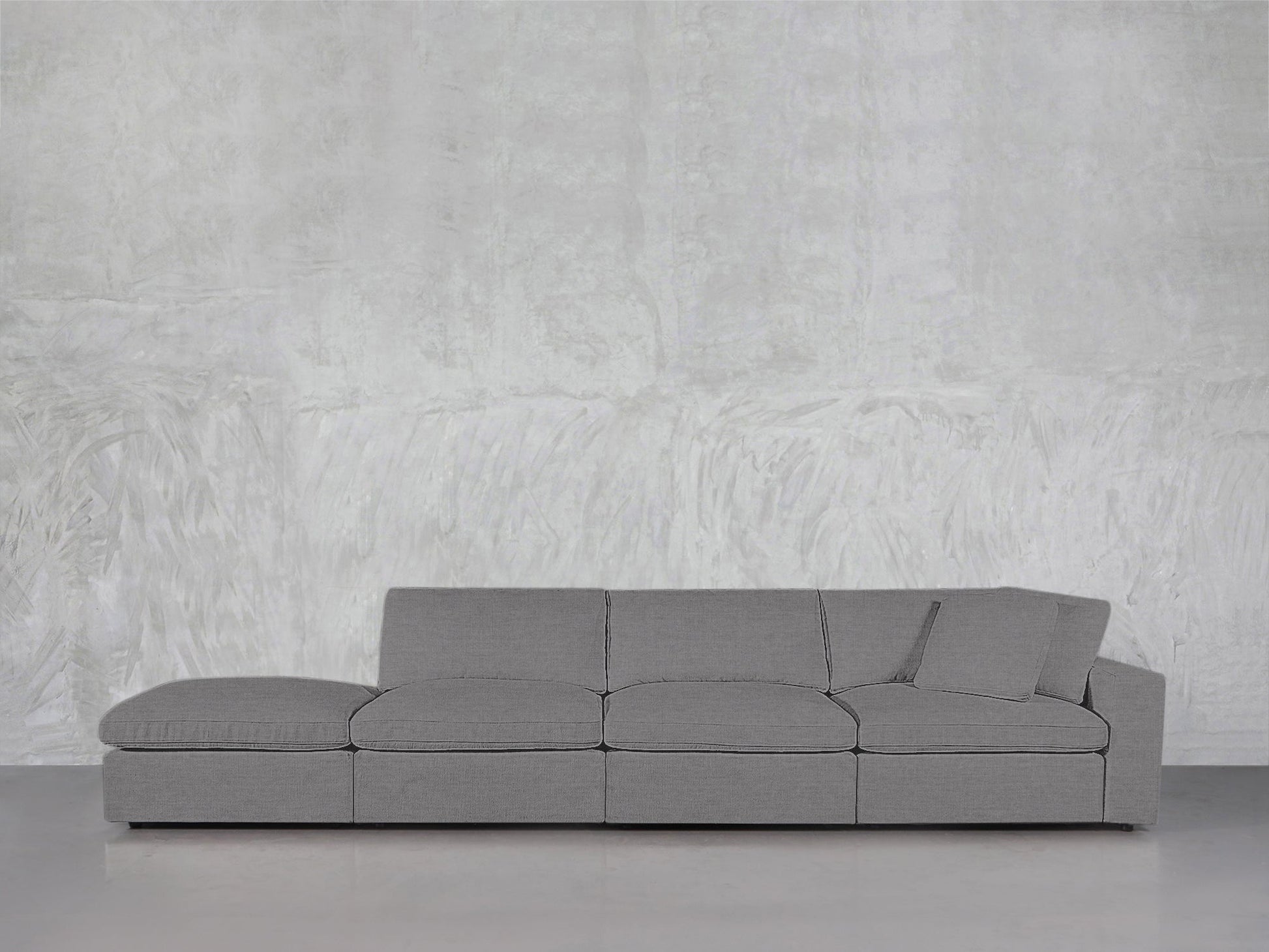 A modern gray 4-Seat Modular Lounger Sofa with matching pillow is arranged against a textured light gray wall and a smooth floor.
