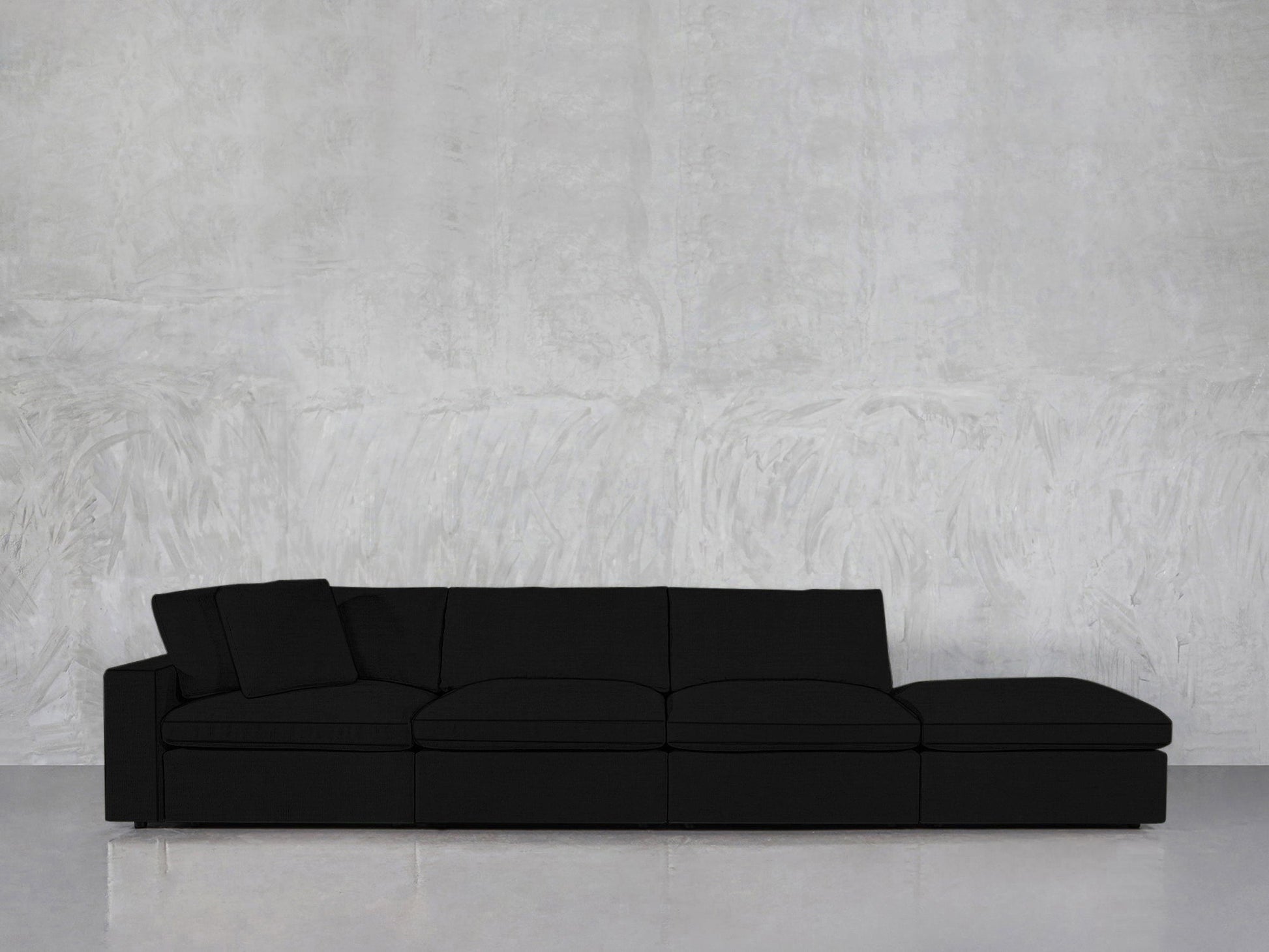 A 4-Seat Modular Lounger Sofa in black with clean lines and a matching cushion sits against a textured light gray wall on a polished floor.