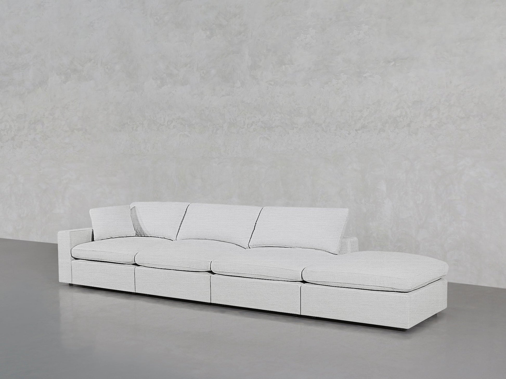 The 4-Seat Modular Lounger Sofa in light gray, featuring four seat and back cushions, sits on a smooth gray floor against a textured pale gray wall.