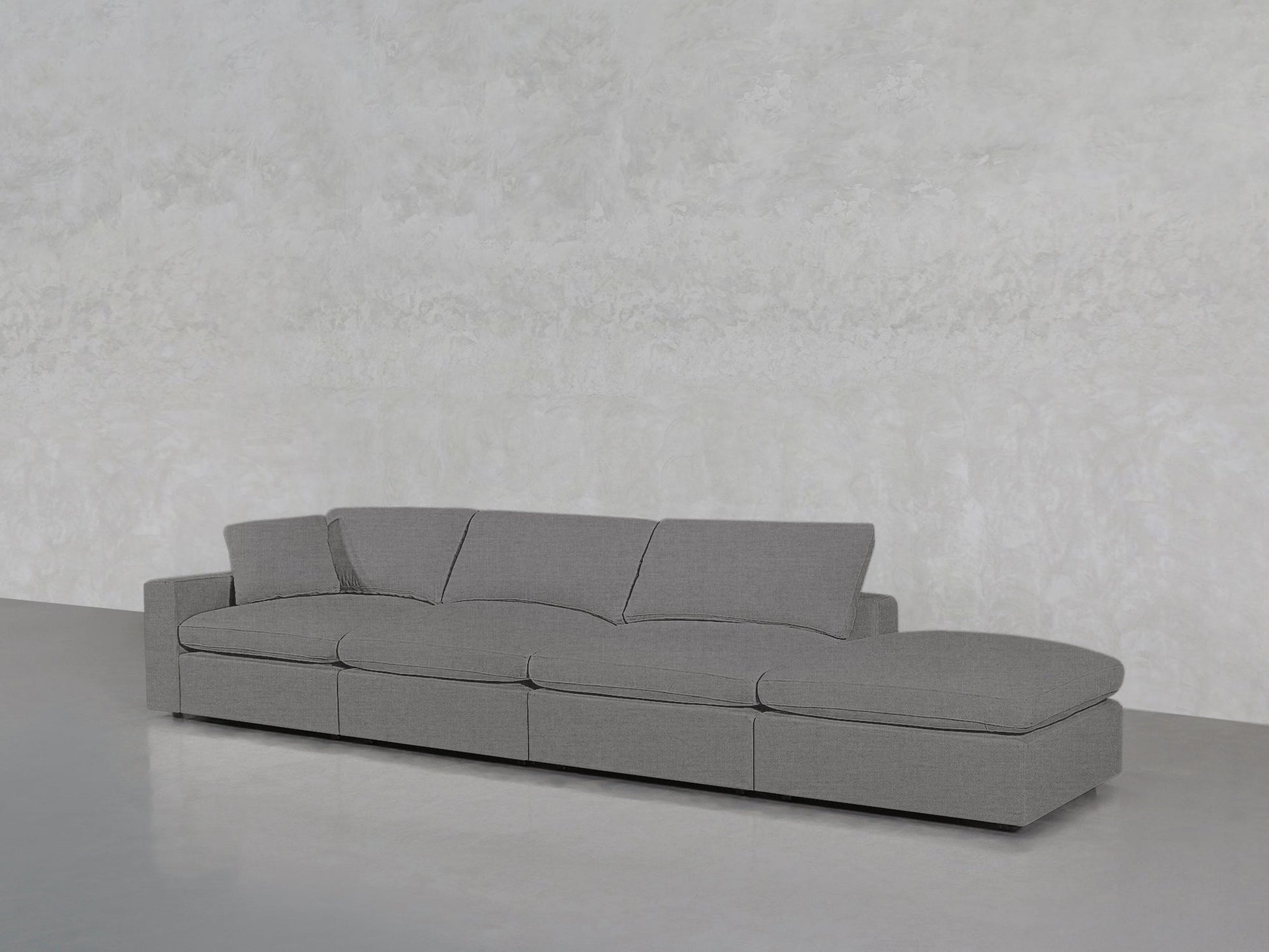 The 4-Seat Modular Lounger Sofa features a modern grey design with three seat cushions and a right-side extended chaise, set against a light grey textured wall and smooth grey flooring.