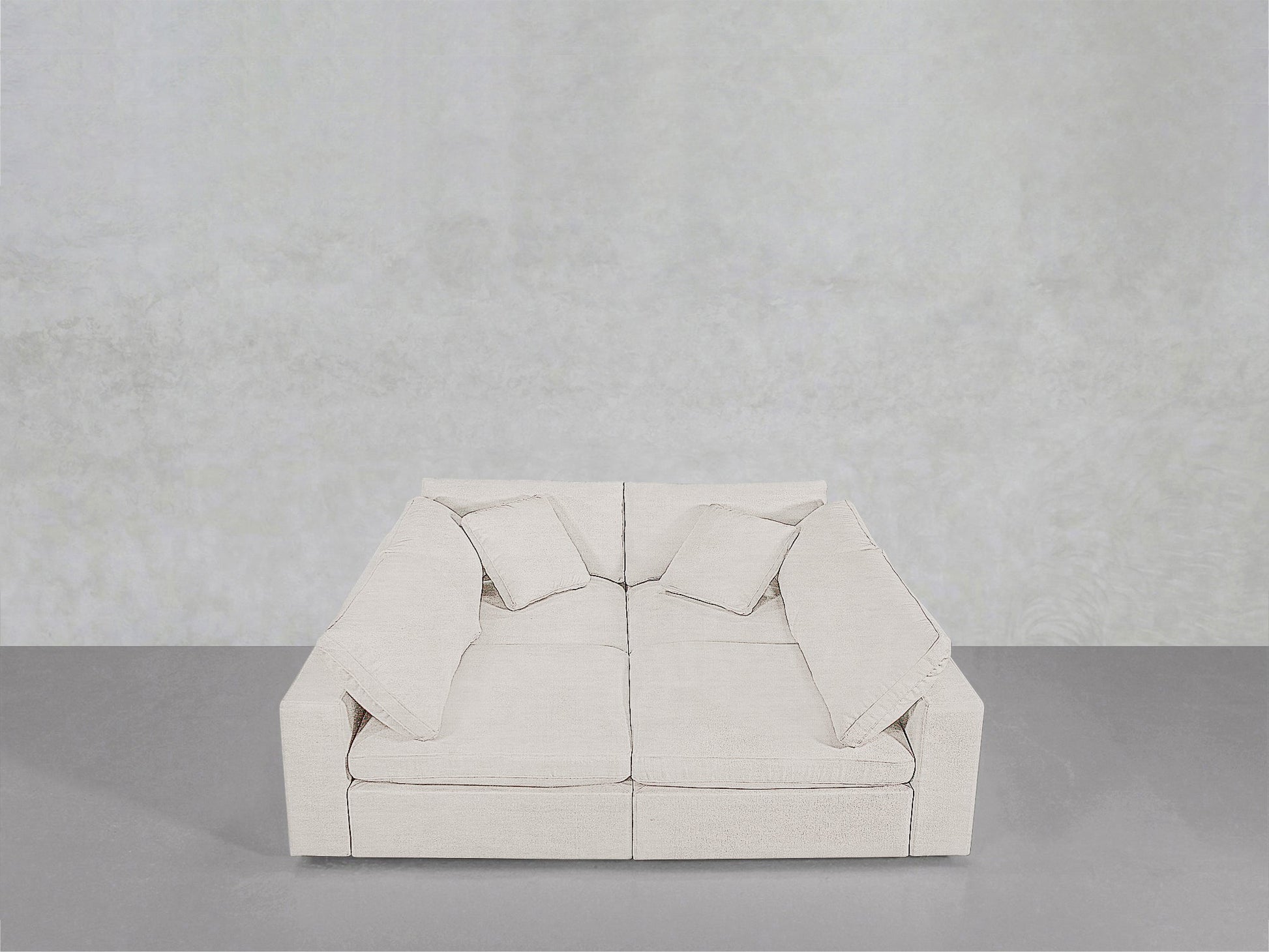 The 4-Seat Modular Pit, a modern light beige sofa with four matching throw pillows, features a minimalist boxy design and clean lines. It sits against a plain light gray wall and gray floor.