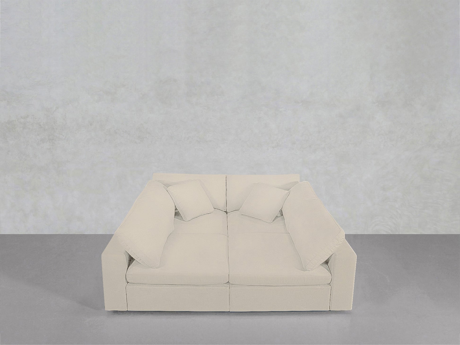 A modern, light beige 4-Seat Modular Pit with four matching cushions is set against a plain, light gray wall on a smooth gray floor.