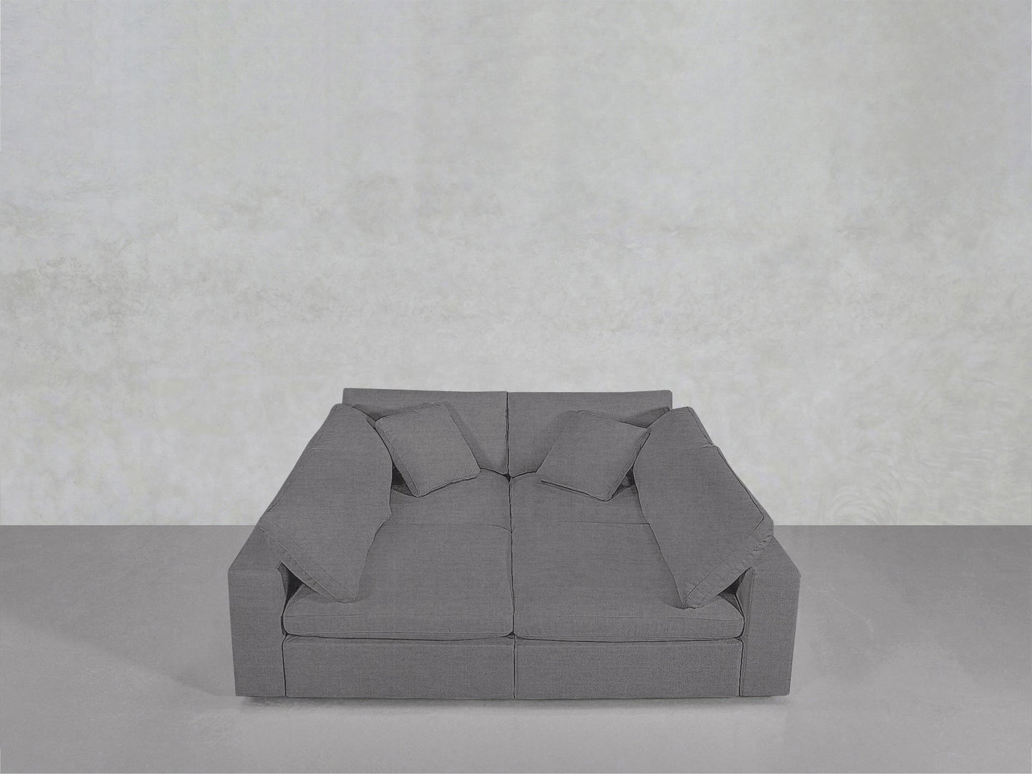 A sleek grey 4-Seat Modular Pit with wide armrests and four matching throw pillows rests against a light grey wall and floor in a minimalist space.
