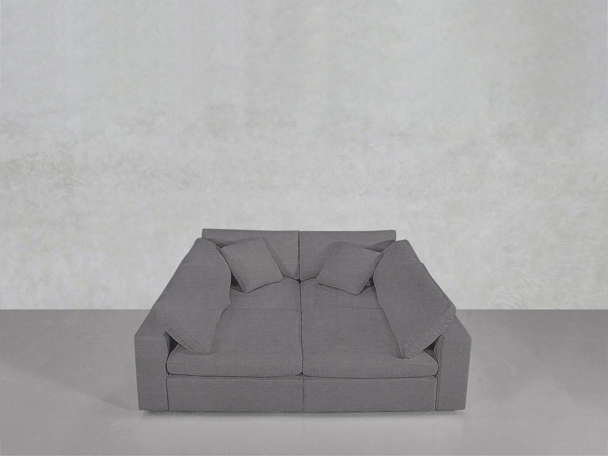 A sleek grey 4-Seat Modular Pit with wide armrests and four matching throw pillows rests against a light grey wall and floor in a minimalist space.