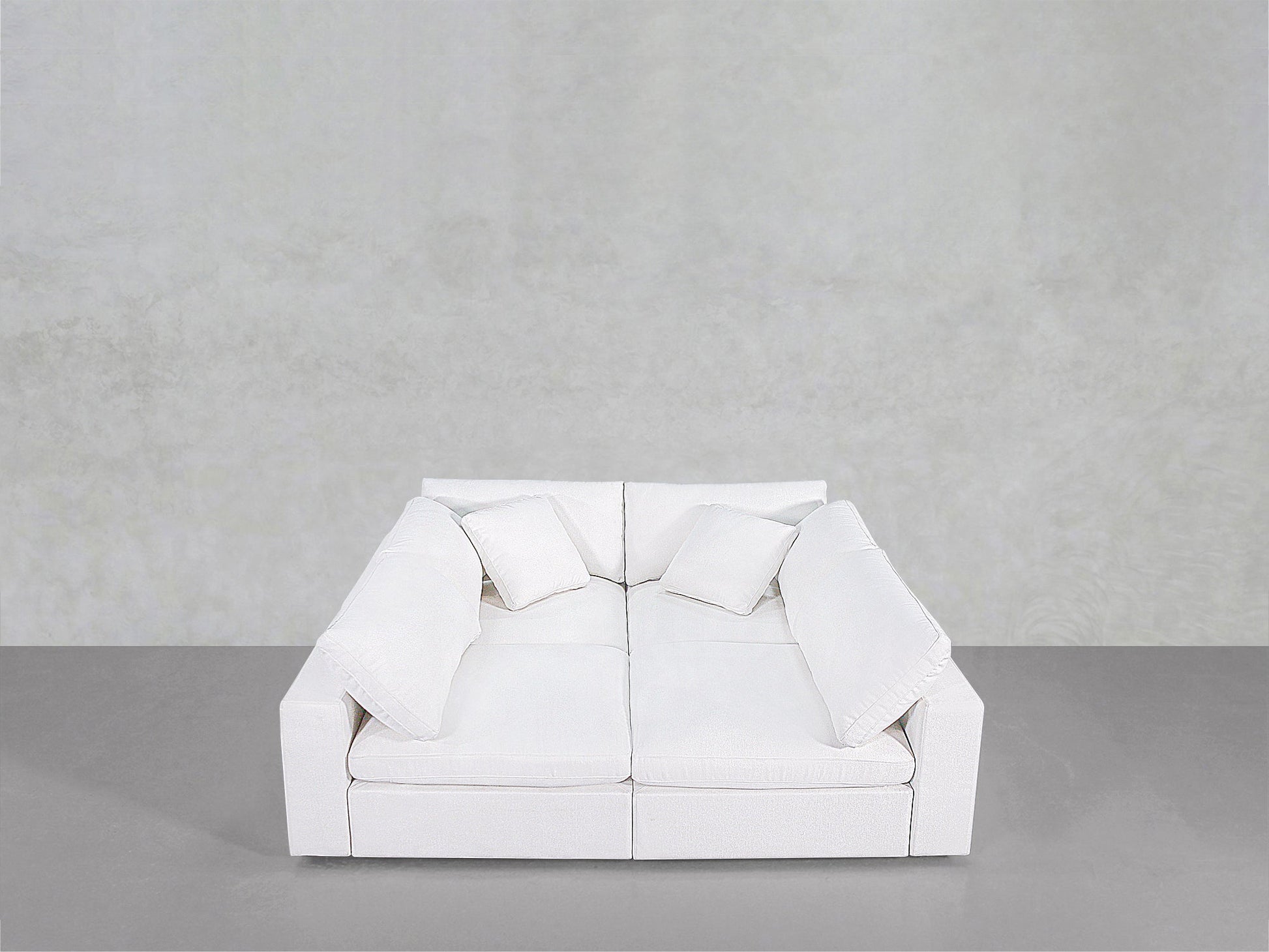 The 4-Seat Modular Pit is a modern white sofa featuring two seat cushions, two back cushions, and four square pillows, shown against a plain light gray wall and gray floor.