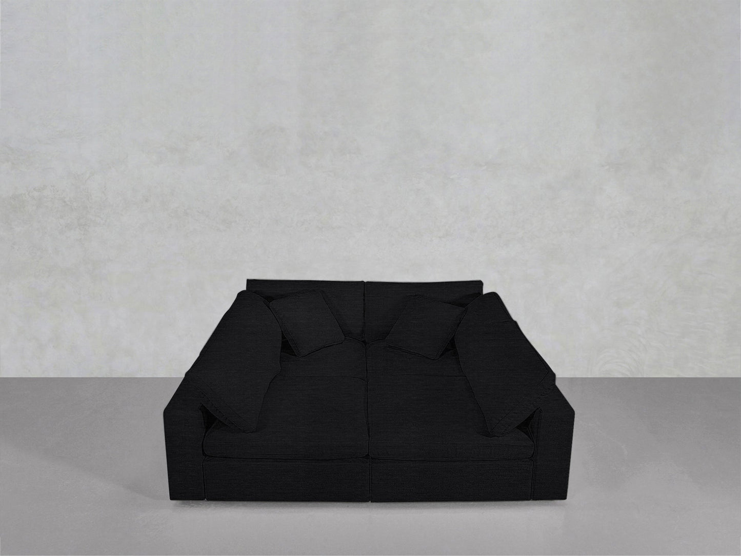 The 4-Seat Modular Pit, a modern dark sofa with four matching cushions, is centered against a plain light gray wall and floor in a minimalist space with no other furniture or decor.