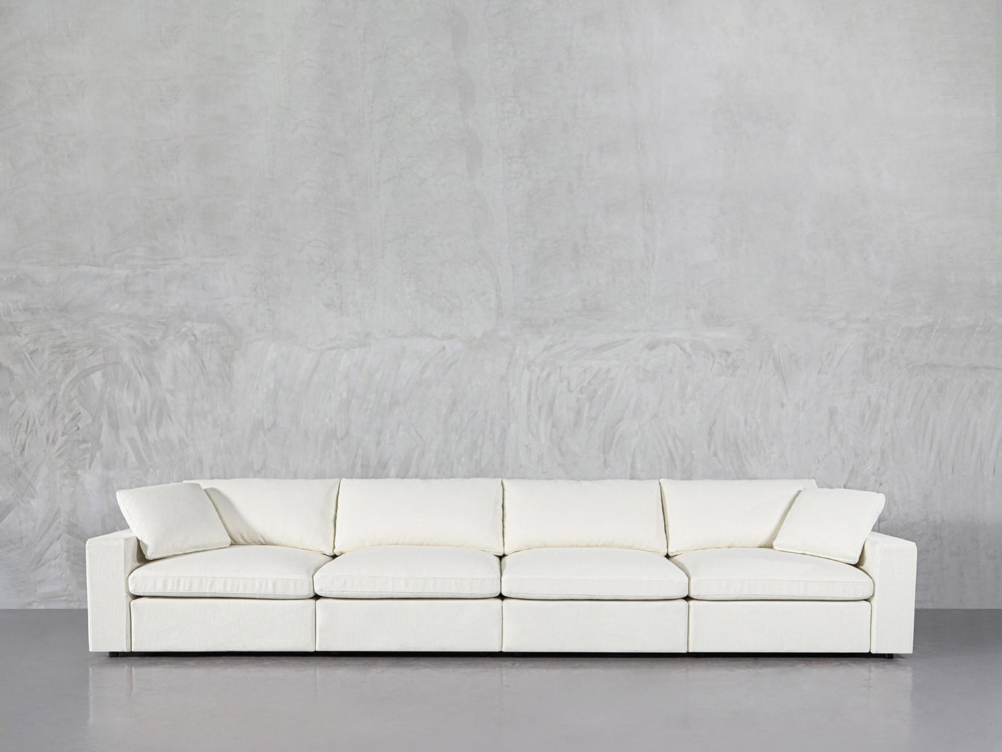 The 4-Seat Modular Sofa, featuring a modern minimalist design with square arms and a low backrest, sits against a light gray textured wall on a polished gray floor.