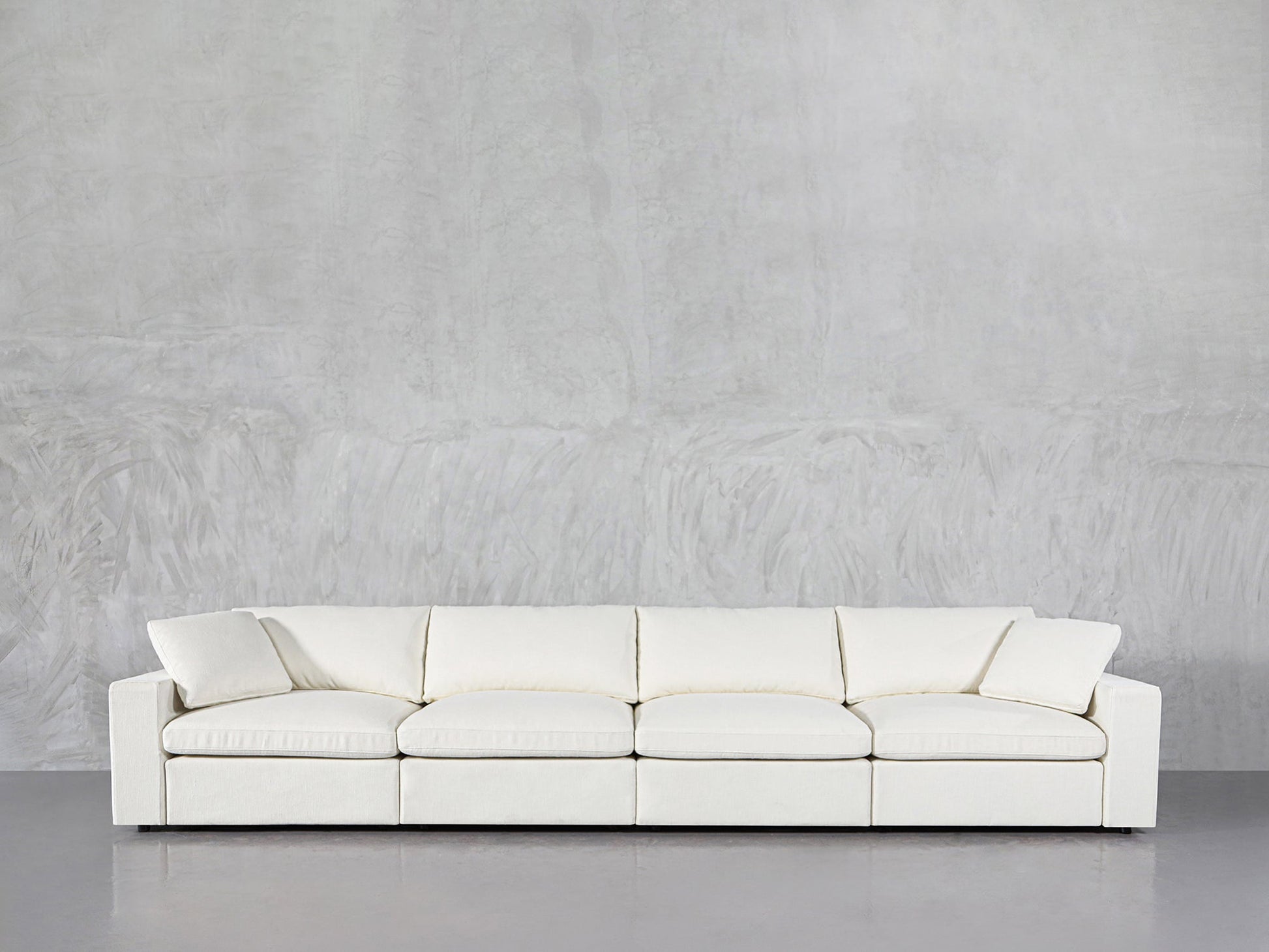 The 4-Seat Modular Sofa, featuring a modern minimalist design with square arms and a low backrest, sits against a light gray textured wall on a polished gray floor.