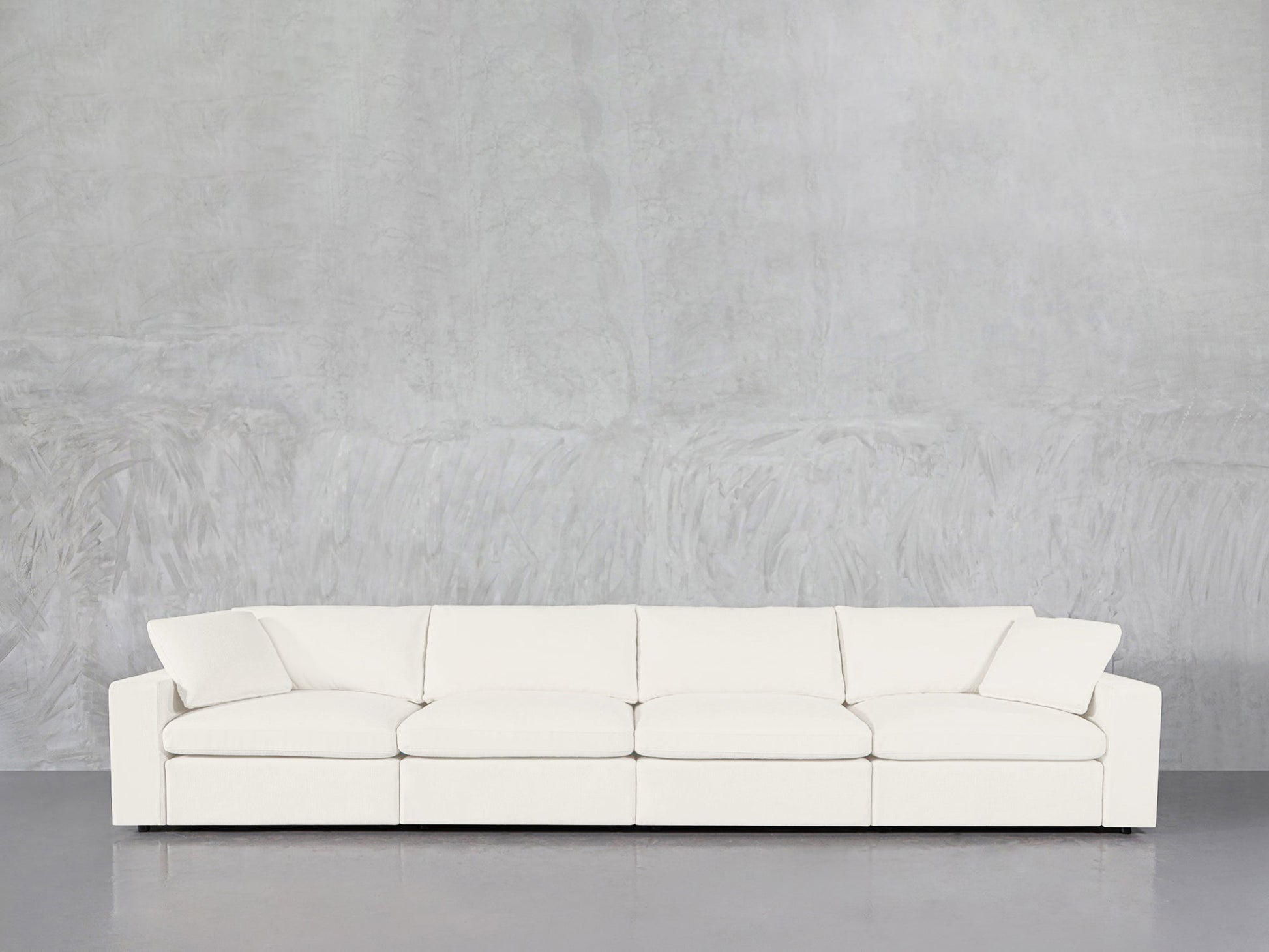 The 4-Seat Modular Sofa, featuring a modern white design and two angled armrests, is arranged against a light gray textured wall and a smooth gray floor.