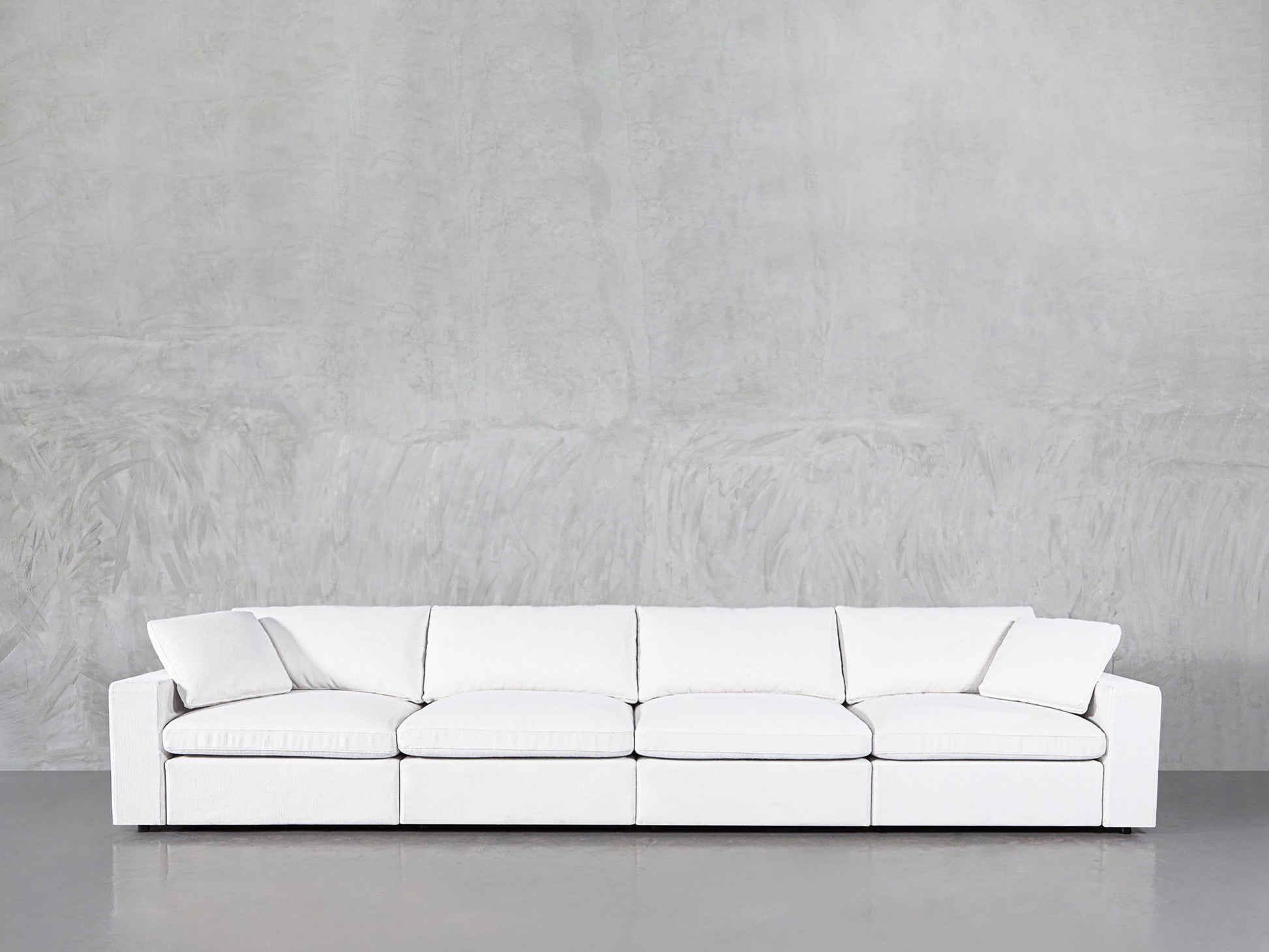 The 4-Seat Modular Sofa, featuring a modern white design with clean lines, is placed against a light gray textured wall and concrete floor.