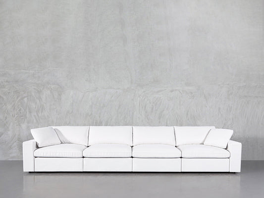 The 4-Seat Modular Sofa, featuring a modern white design with clean lines, is placed against a light gray textured wall and concrete floor.