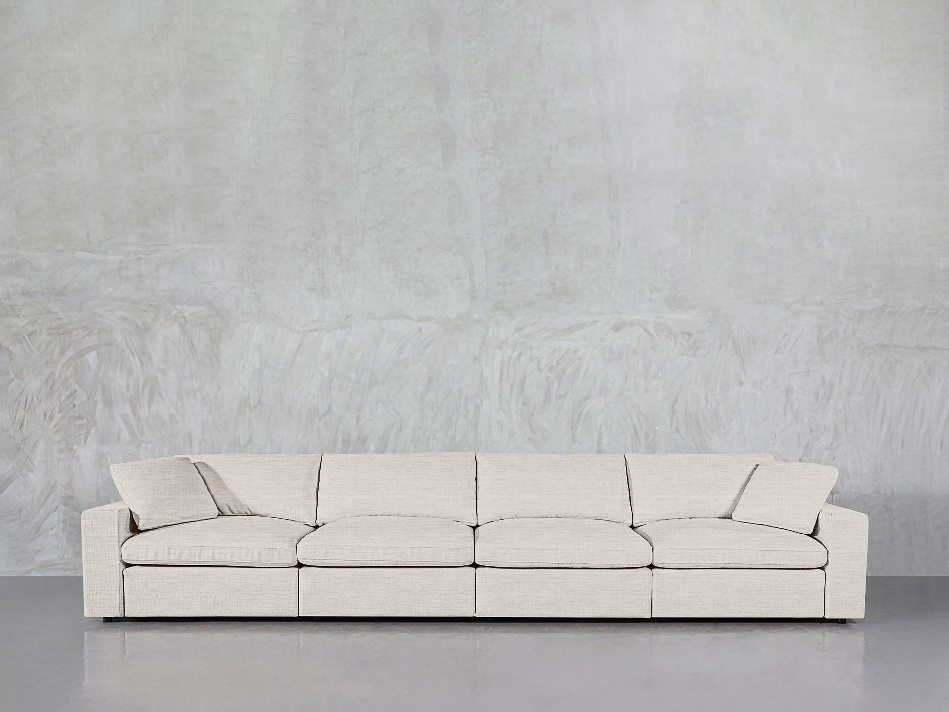 A modern 4-Seat Modular Sofa in light beige with matching pillows is placed against a textured light gray wall on a reflective gray floor.