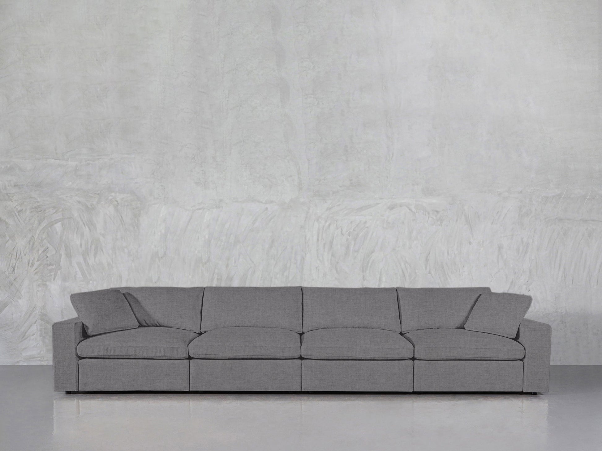 The 4-Seat Modular Sofa, featuring a modern minimalist design with four seat cushions and two armrests, is positioned against a light grey textured wall on a polished floor.