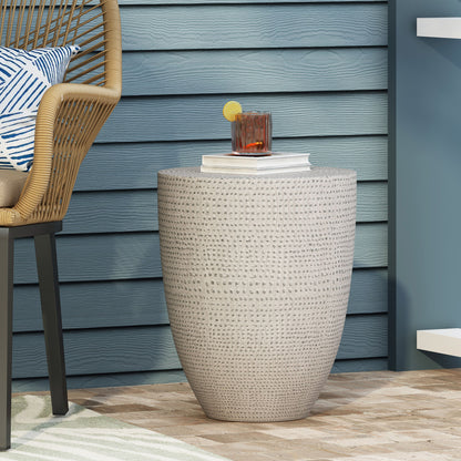 The Outdoor Lightweight Concrete Side Table in Natural sits on a patio with a blue wooden wall, holding a drink and book; part of a wicker chair is visible on the left.