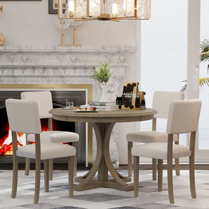 A Retro Round Dining Table Set with curved trestle legs and upholstered chairs is placed in front of a marble fireplace, topped with a vase, books, and a card game set. A chandelier hangs above, and large windows fill the background.
