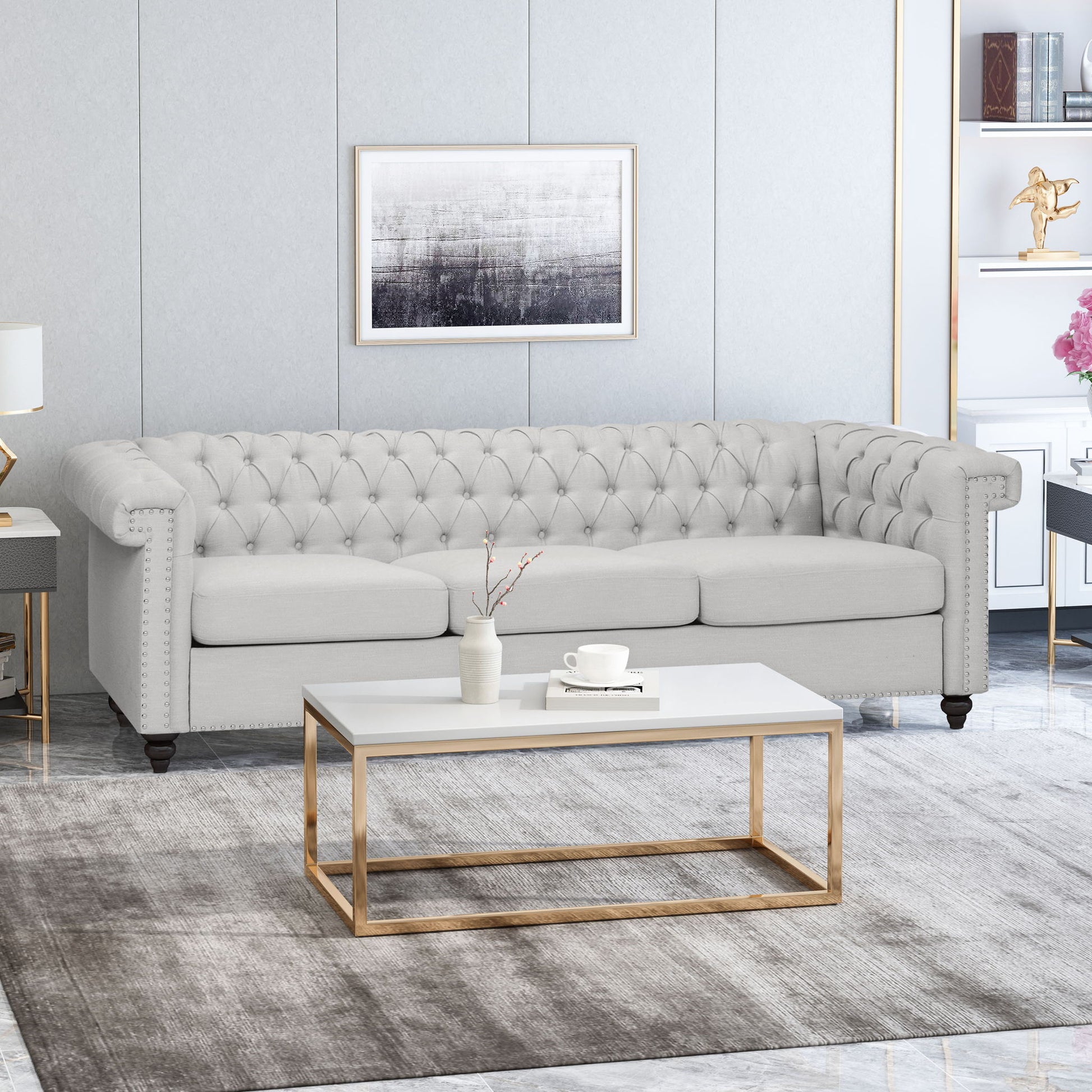 A modern living room features a Chesterfield Style 3 Seater Sofa With Deep Button Tufting in light gray, a white and gold coffee table with a mug and vase, a gray rug, and abstract wall art above the sofa for an elegant neutral look.
