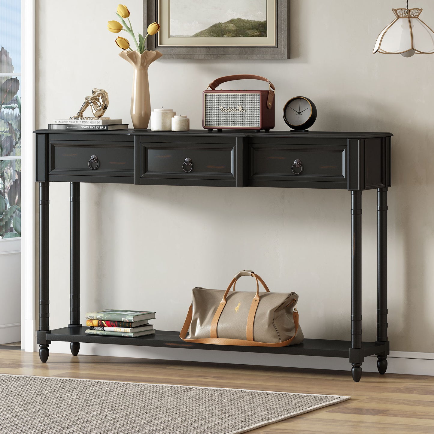 The Console Table Sofa Table With Drawers For Entryway features three projecting drawers and a long lower shelf, topped with a vase of yellow tulips, books, a vintage radio, and sculpture. A tan duffel bag and stack of books are on the bottom shelf.