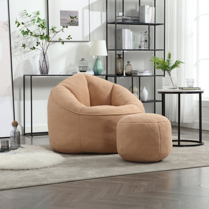 The Bedding Bean Bag Sofa Chair High Pressure Foam With Footrest sits on a beige rug in a modern living room with plants, shelves, books, and décor. Sunlight streams through large windows, highlighting its cozy tan design.