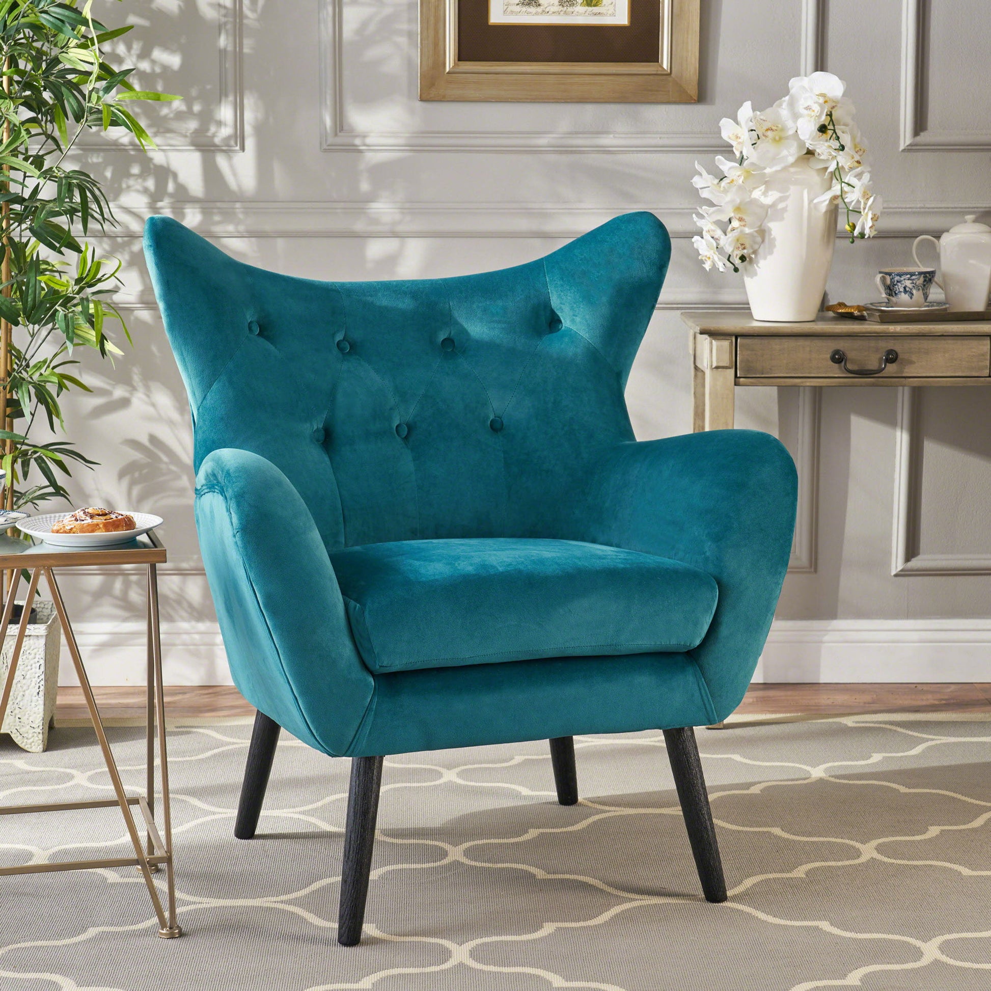 The Alyssa Mid Century Upholstered Arm Chair with wingback design, featuring teal velvet and black tapered legs, sits on a light patterned rug next to a side table with a pastry, potted plant, and a console adorned with a white orchid.