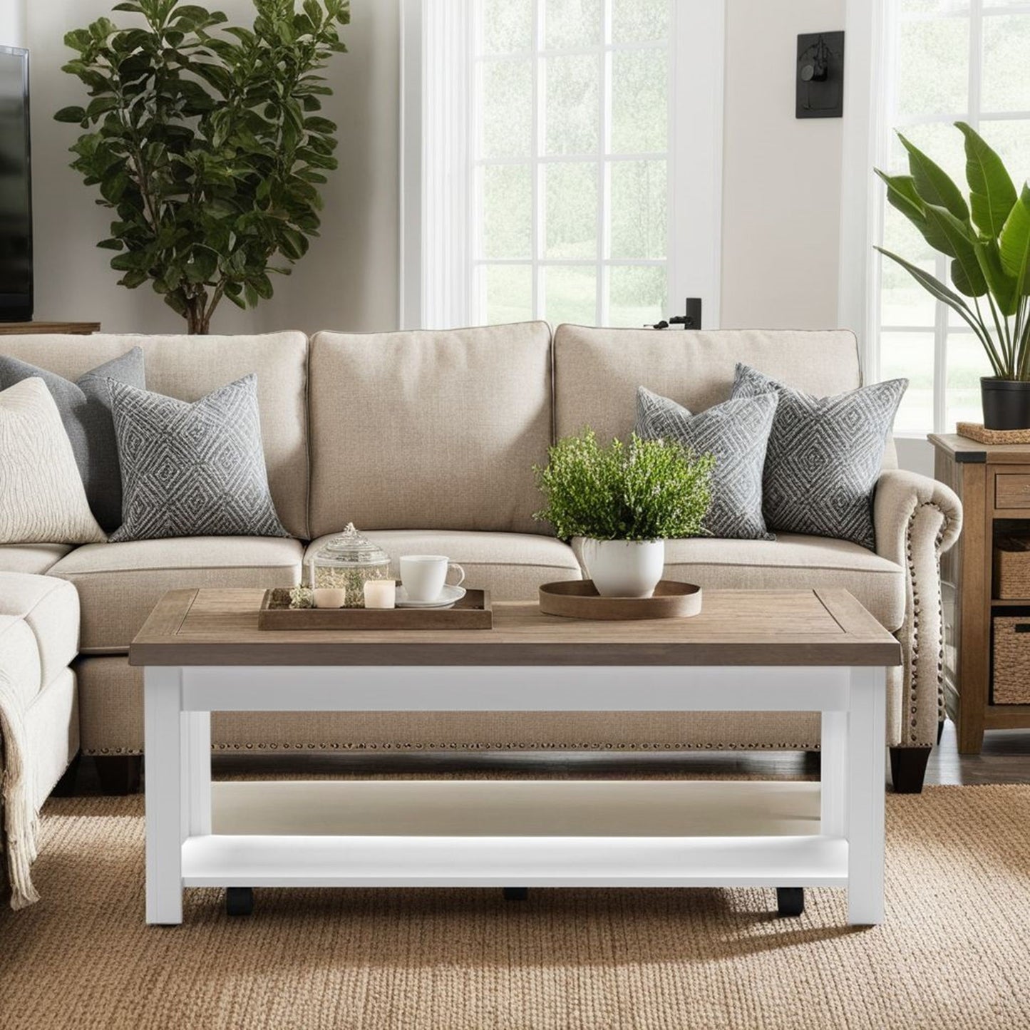 Neutral-toned living room with a beige sofa, gray patterned pillows, the Hampton Coffee Table in Jasmine Whitewash/Barnwood displayed with a plant, cup, and tray, a jute rug, and two large potted plants by a bright window with white trim.