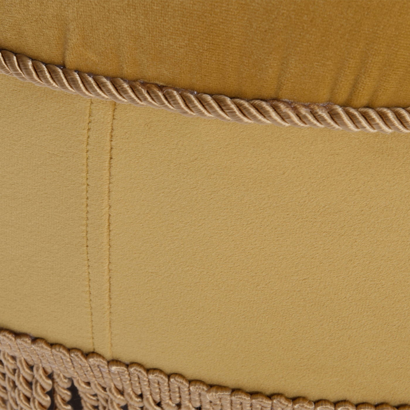 Close-up of the Yolanda - Luxurious Upholstery Round Upholstered Accent Ottoman in mustard yellow fabric, featuring a stitched seam, twisted cord trim, and fringed trim along the bottom edge.