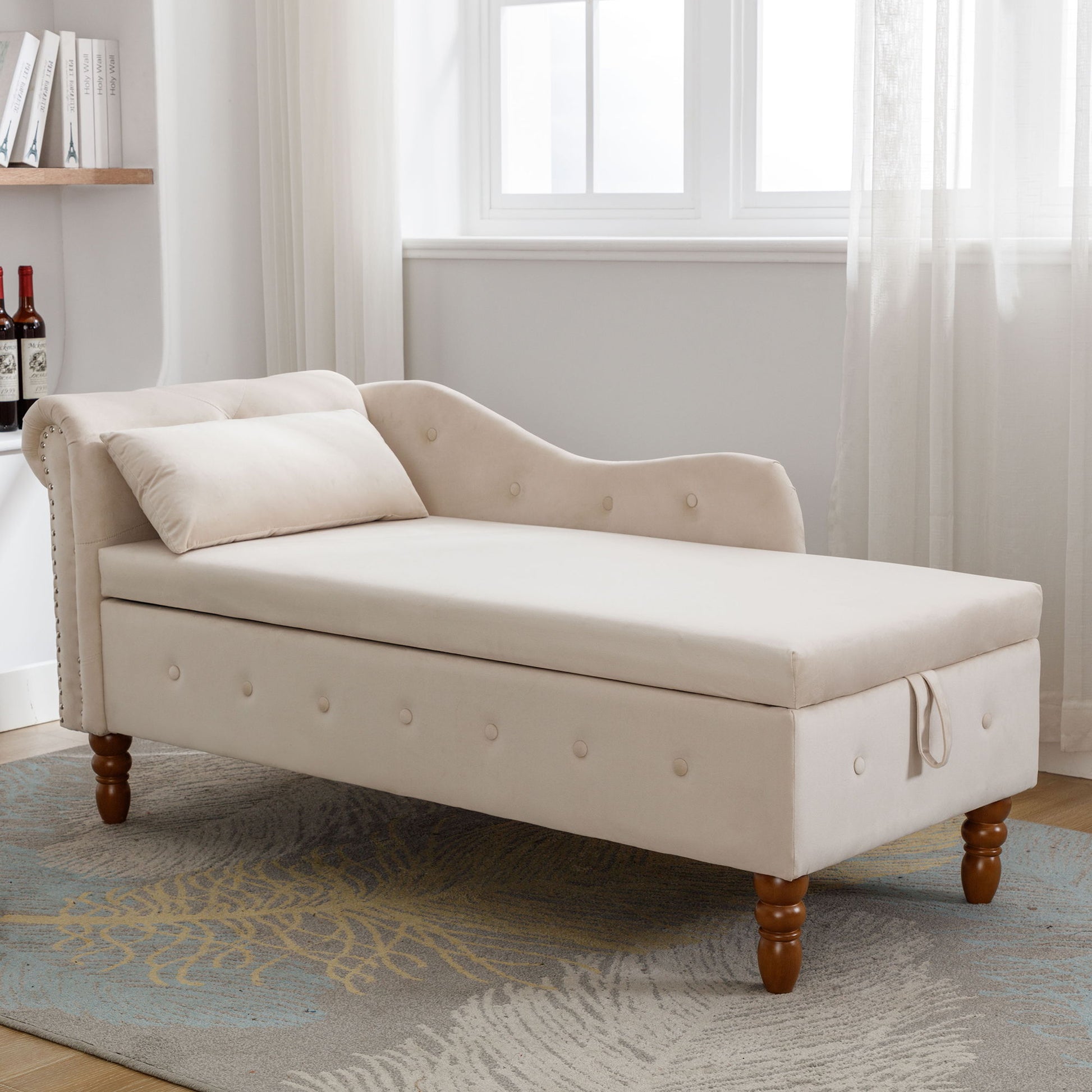 A modern beige velvet chaise lounge with rolled arms, nailhead trim, storage, and a matching pillow sits on a patterned rug by a window with sheer curtains in a bright room.