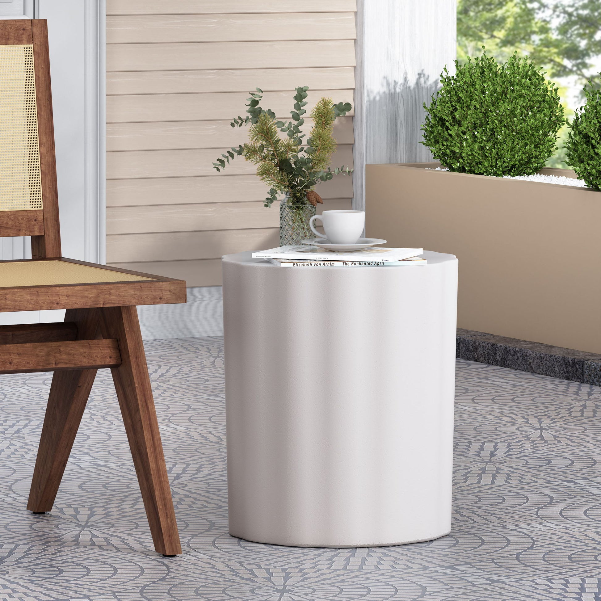 The Outdoor End Table With Soft Wave Edge in Antique White sits beside a wooden chair on patterned tiles, displaying a milk cup, plate, plant, and magazines, with green bushes and a beige wall in the background.
