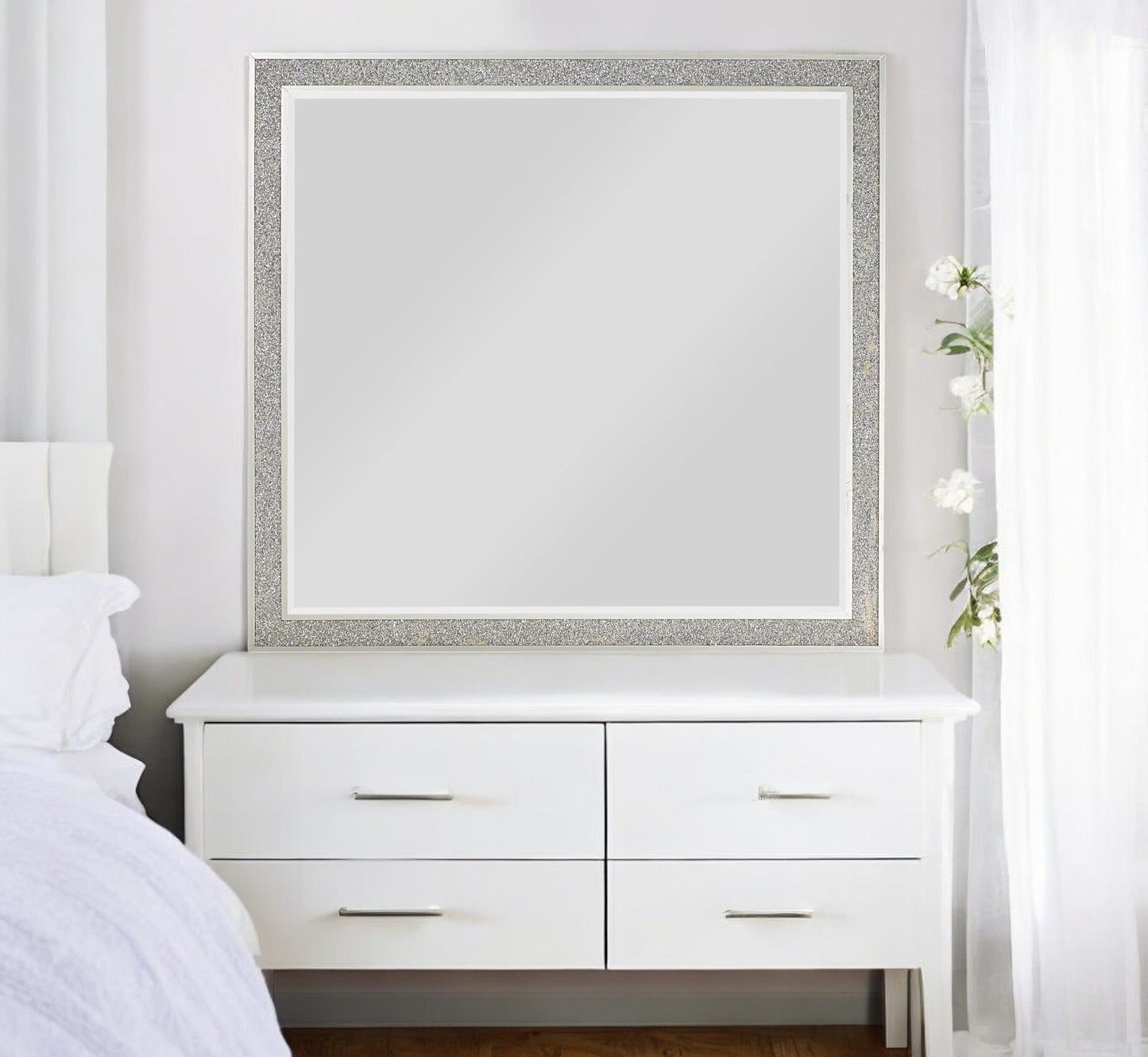 The Sliverfluff Mirror in Mirrored/Champagne hangs above a white four-drawer dresser; the room features white walls, bedding, sheer curtains, and a tall vase of white flowers to the right.