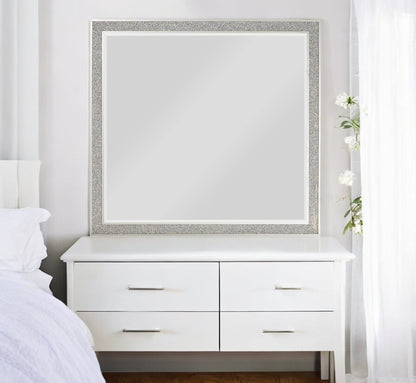 The Sliverfluff Mirror in Mirrored/Champagne hangs above a white four-drawer dresser; the room features white walls, bedding, sheer curtains, and a tall vase of white flowers to the right.