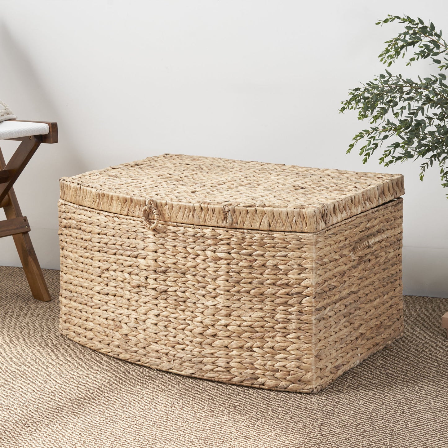 The Rectangular Curve Water Hyacinth Woven Wicker Trunk with handles in natural light brown sits on a textured rug near a wooden chair and green plant, perfect for storing clothes, toys, magazines, and books against a white wall.