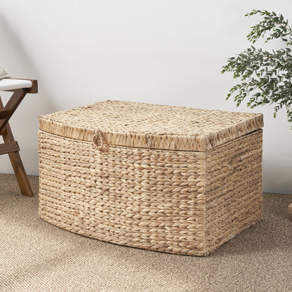 The Rectangular Curve Water Hyacinth Woven Wicker Trunk with handles in natural light brown sits on a textured rug near a wooden chair and green plant, perfect for storing clothes, toys, magazines, and books against a white wall.