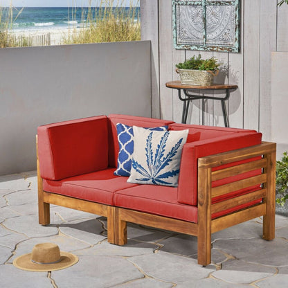 The Oana 2 Piece Loveseat Set with water-resistant cushions sits on a stone patio near the beach, accented by red cushions, two blue-and-white pillows, and a straw hat, with coastal grasses and ocean in the background.