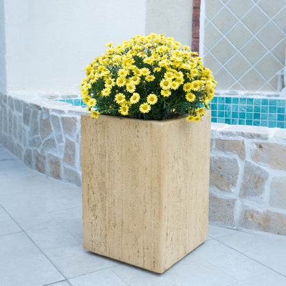 The Modern Outdoor Planter, a square beige stone design filled with yellow daisies, sits on a tiled patio near a stone wall and blue-tiled pool.