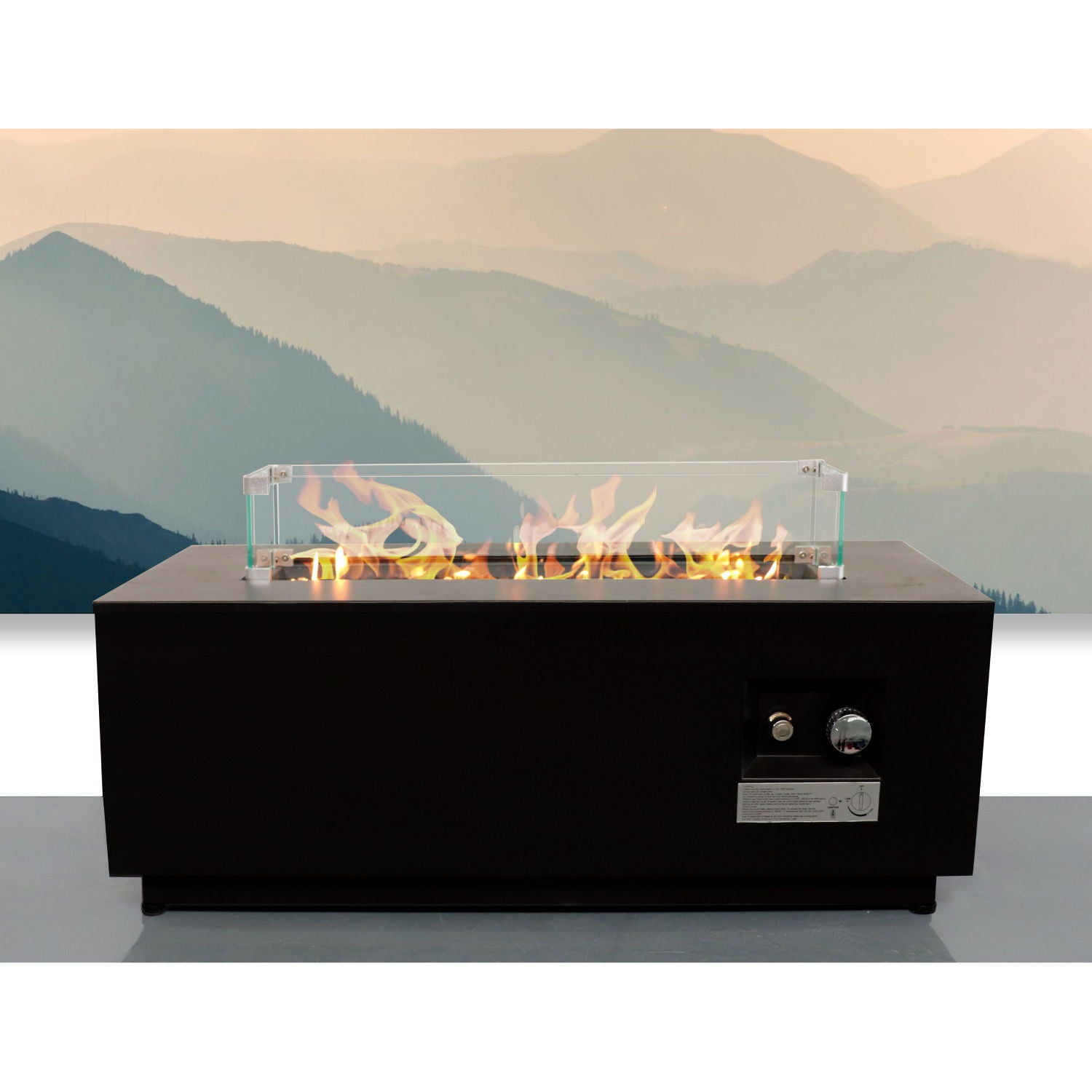 A black Outdoor Fire Pit Table With Lid, featuring fuel versatility, glass wind guard, visible flames, and side control knobs, is set before hazy blue mountains.