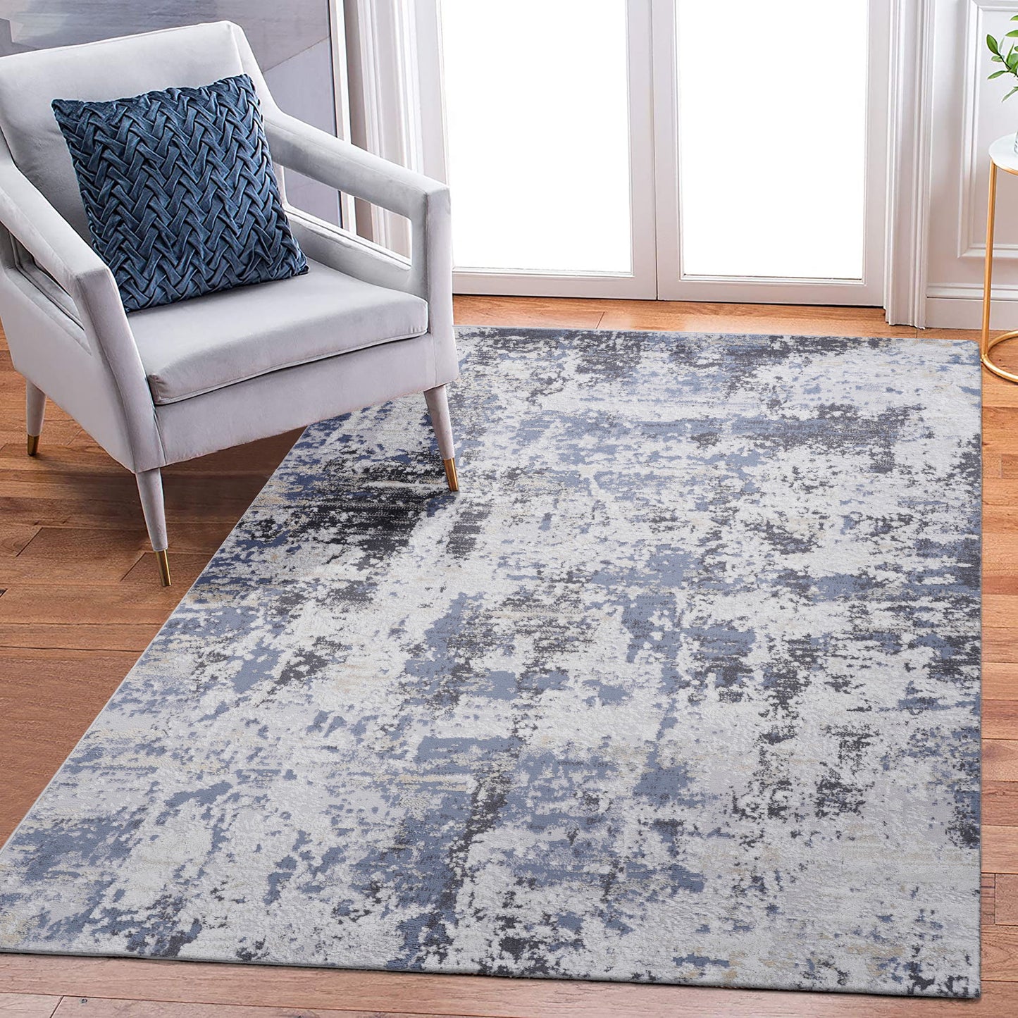 A modern living room features a light gray armchair with a blue patterned cushion and the Payas - Abstract Area Rug in blue, gray, and white on a wooden floor near glass doors.