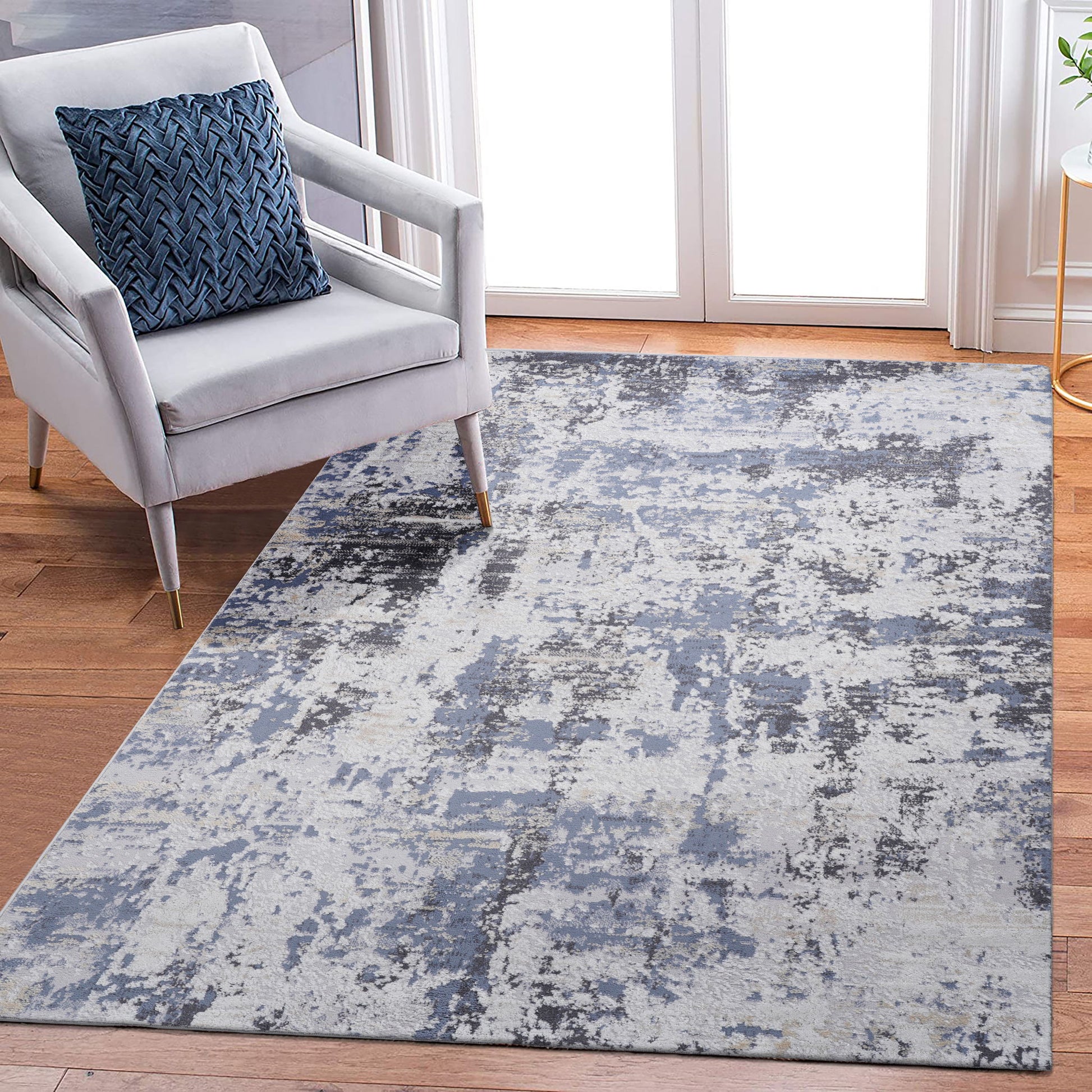 A modern living room features a light gray armchair with a blue patterned cushion and the Payas - Abstract Area Rug in blue, gray, and white on a wooden floor near glass doors.