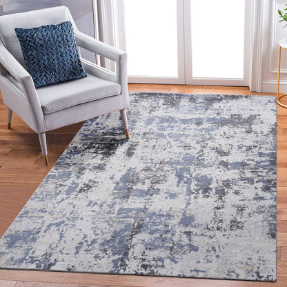 A modern living room features a light gray armchair with a blue patterned cushion and the Payas - Abstract Area Rug in blue, gray, and white on a wooden floor near glass doors.