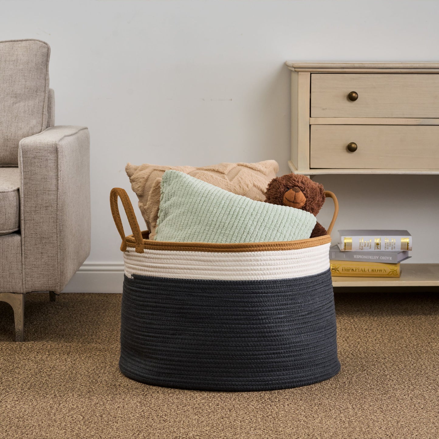 The Francis - Cotton Round Laundry And Storage Basket with tan handles holds two textured pillows and a brown teddy bear. It rests on a carpeted floor beside a beige sofa and a light wood dresser topped with books and a candle.