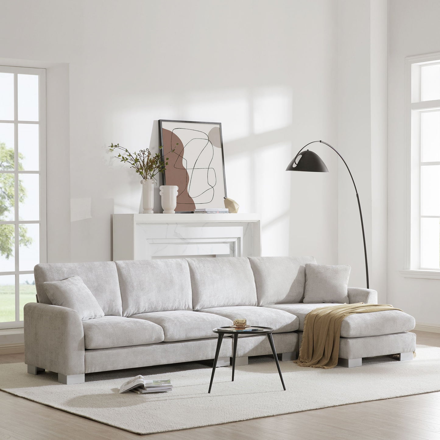 A modern living room features the Modern Oversized Sectional Sofa, an L-shaped chenille couch set with chaise and 2 free pillows, paired with a beige throw, black round coffee table, floor lamp, abstract art above a white fireplace, and large windows.