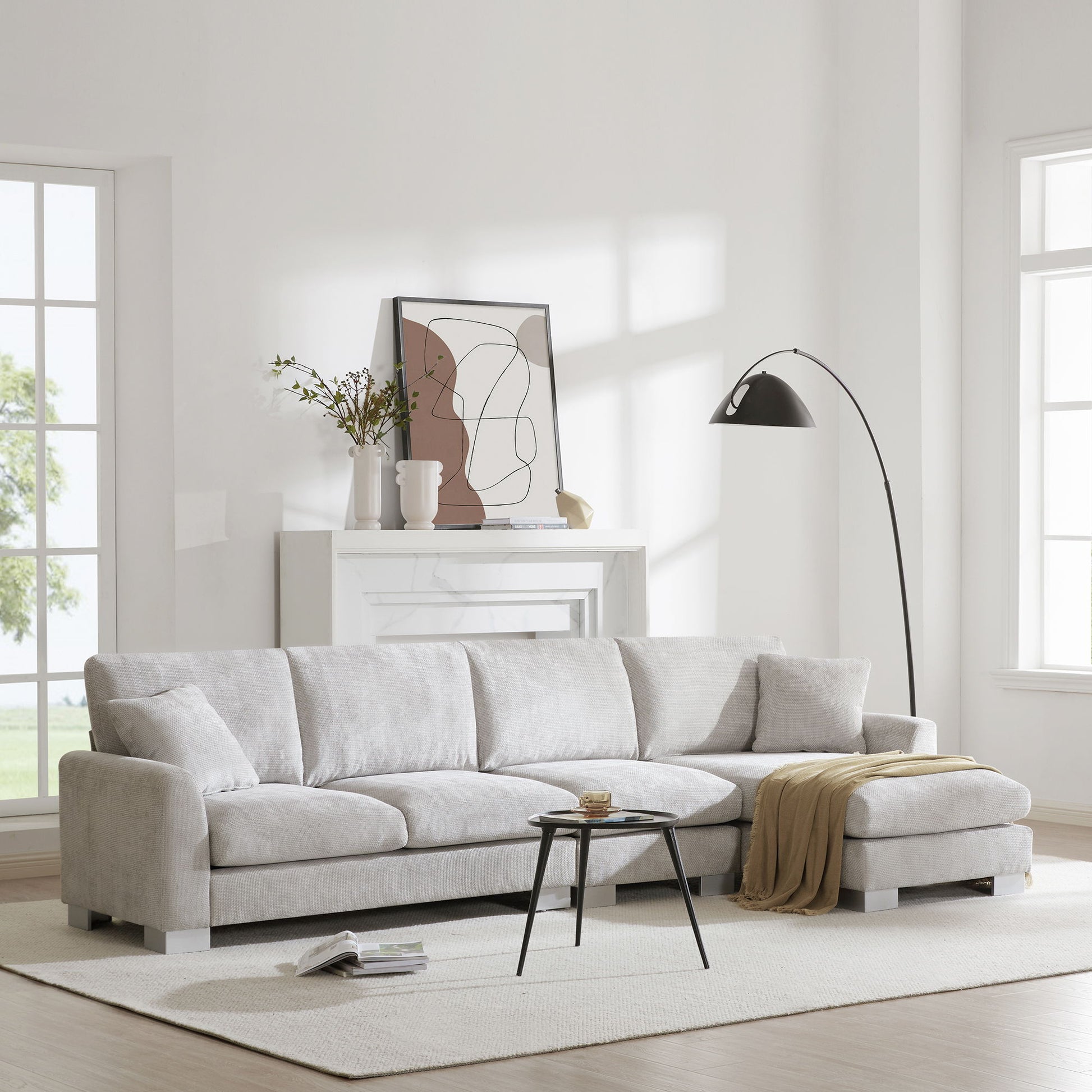 A modern living room features the Modern Oversized Sectional Sofa, an L-shaped chenille couch set with chaise and 2 free pillows, paired with a beige throw, black round coffee table, floor lamp, abstract art above a white fireplace, and large windows.